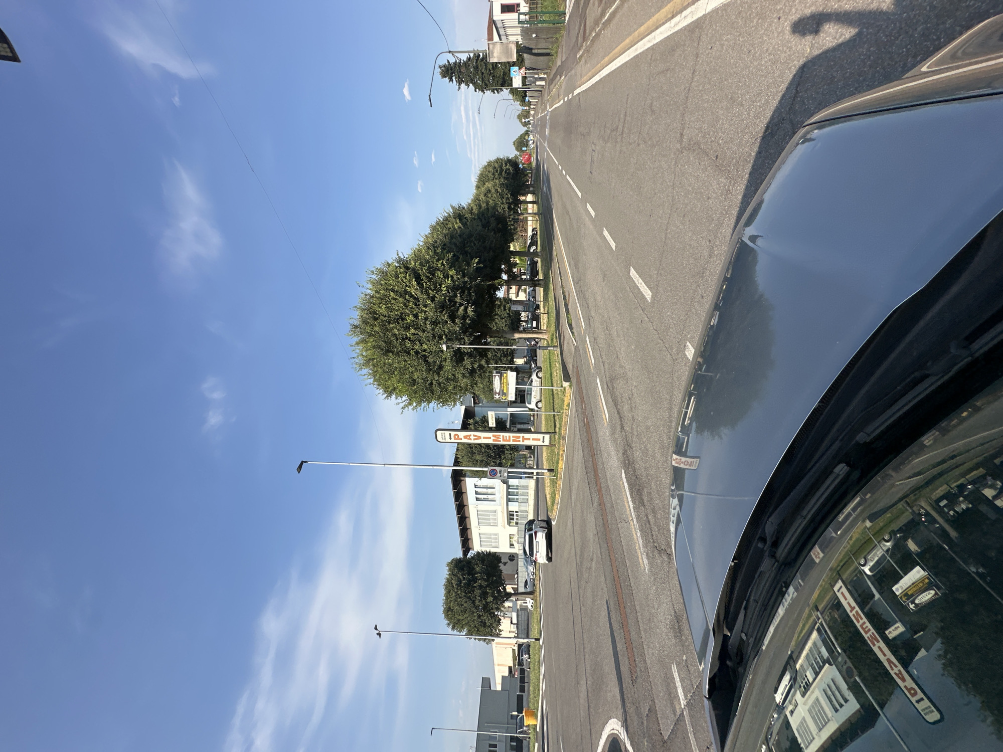 The image shows a street scene in Rudiano, Italy, viewed from the perspective of someone inside a car. The car's hood and windshield are visible in the foreground, reflecting the surrounding environment. The street is paved with asphalt and marked with white lines indicating traffic lanes. On the left side of the street, there is a roundabout with a yellow bollard. Further down the street, there are buildings, trees, and streetlights. Several cars are parked or driving on the street. A tall vertical sign on the right side of the road reads "PAVIMENTI" with an arrow pointing to the right, indicating flooring or pavement services. The sky is blue with scattered white clouds. The overall scene depicts a typical urban or suburban environment during the daytime.