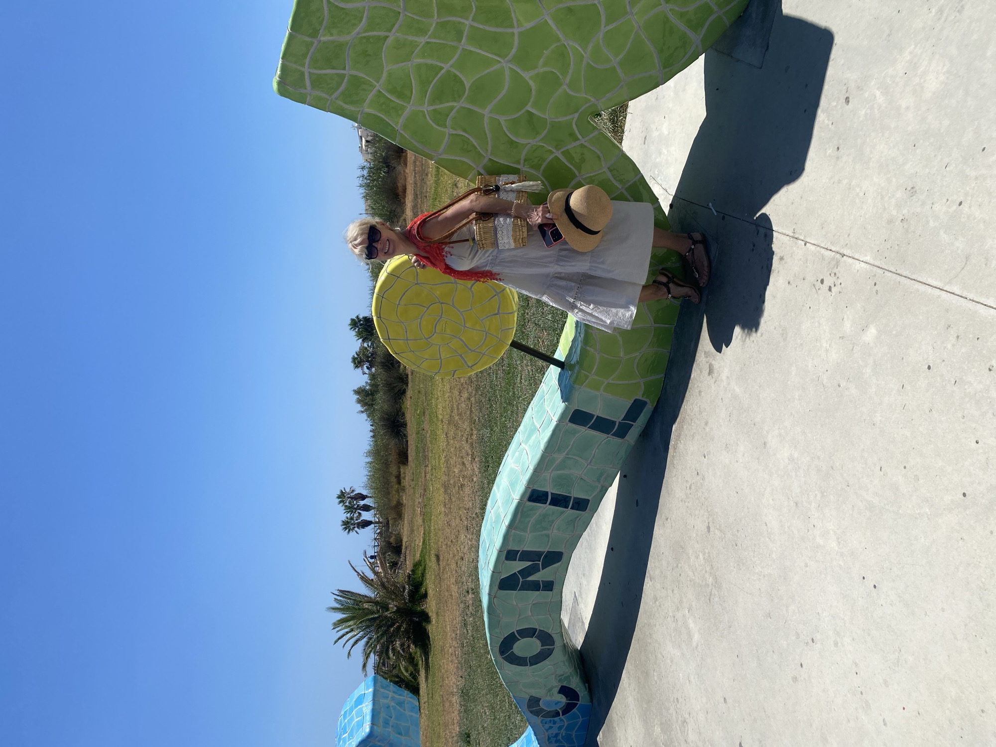 A woman with blonde hair and sunglasses, wearing a white sundress, a red scarf, and sandals, poses for a photo in front of a large, mosaic-tiled art installation in Conil De La Frontera, Spain. The art piece spells out "CONIL" and features vibrant blue, green, and yellow tiles. The woman is holding a straw bag and a straw hat. The background consists of a grassy field, palm trees, and a clear blue sky. The ground is paved with concrete.