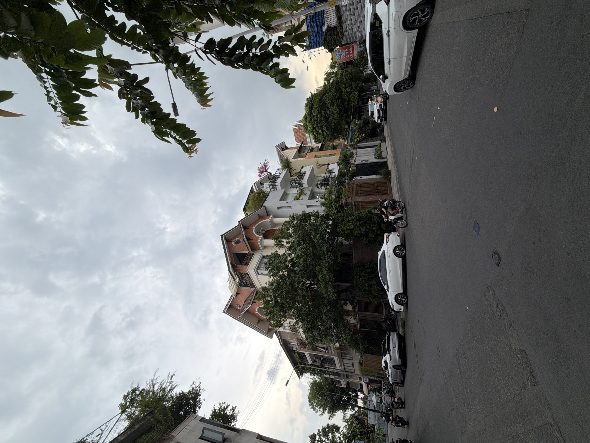 The image depicts a street scene in Thủ Đức City, Vietnam, with a focus on a multi-story building and the surrounding urban environment. The sky is overcast with dense clouds.
On the right side of the street, a white SUV is parked, its rear section facing the camera. Further down the street, another white car, a sedan, is parked. In the foreground, a silver Porsche is also parked. Several motorcycles are present, with some riders visible.
The building on the left is a tall, multi-story structure with a distinctive architectural style, featuring multiple balconies, large windows, and decorative elements like arched openings and red-tiled rooflines. Lush green trees and foliage surround the building and are integrated into its design, with plants on balconies and a large tree in front. A street light is visible near the building.
The street itself is paved and appears to be a well-trafficked urban road. Signs with Vietnamese text are visible on the side of the road, suggesting commercial activity. The overall impression is a dense urban area with a mix of residential and possibly commercial buildings, characterized by lush greenery.