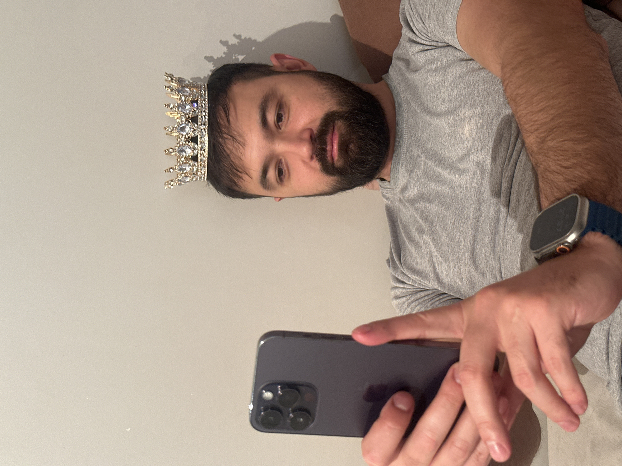 An eye-level selfie shot vertically shows a man in what appears to be an indoor setting, likely a room with a light-colored, plain wall. The man, who has dark hair and a full beard, is looking directly at the camera (his smartphone). He is wearing a grey short-sleeved t-shirt. A golden-colored crown, adorned with clear or white jewels, sits on his head. His expression is neutral.
He holds a dark-colored smartphone, possibly an iPhone given the triple-camera module visible on the back, in his left hand, pointing it towards himself to take the picture. His right arm is bent, and his hand rests on his left forearm/wrist area. On his right wrist, he wears a smartwatch with a blue band and a silver-toned case. A subtle shadow is cast on the wall behind his head, possibly from his hair or the crown. The overall lighting suggests an indoor environment. No discernible text is visible on any object. The location context for this image is Khimki, Russia.