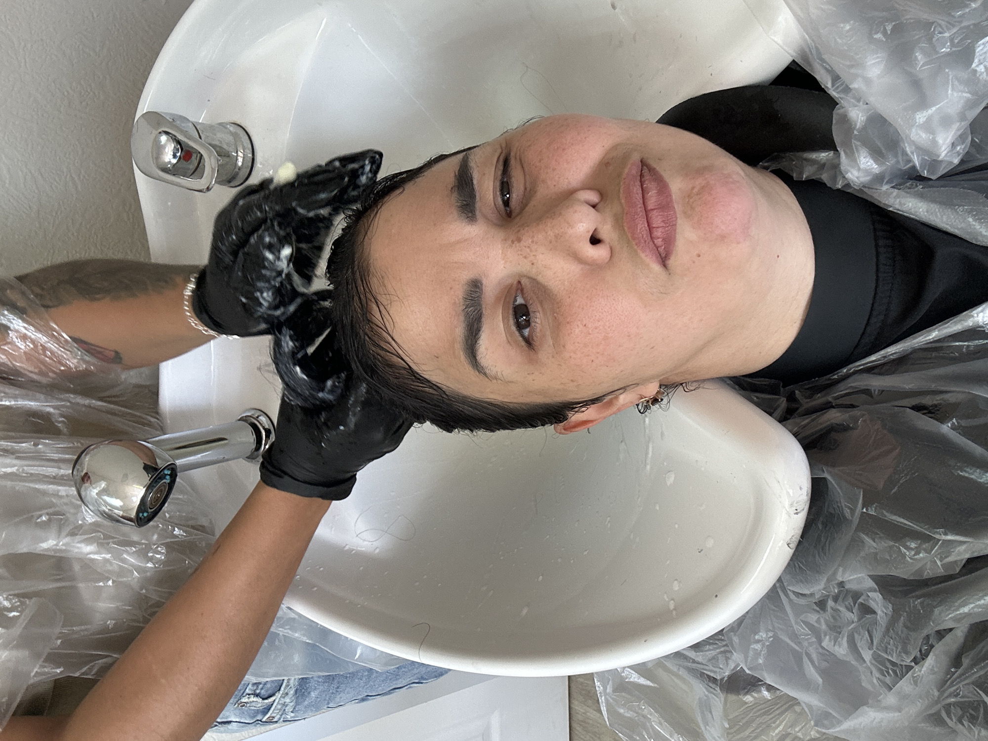 A woman is having her hair washed in a salon sink. The sink is white and oval shaped. The woman has dark hair and light skin. She is wearing a black shirt and plastic wrap. A person with tattooed arms is washing her hair. They are wearing black gloves. The location is Unknown, Unknown.