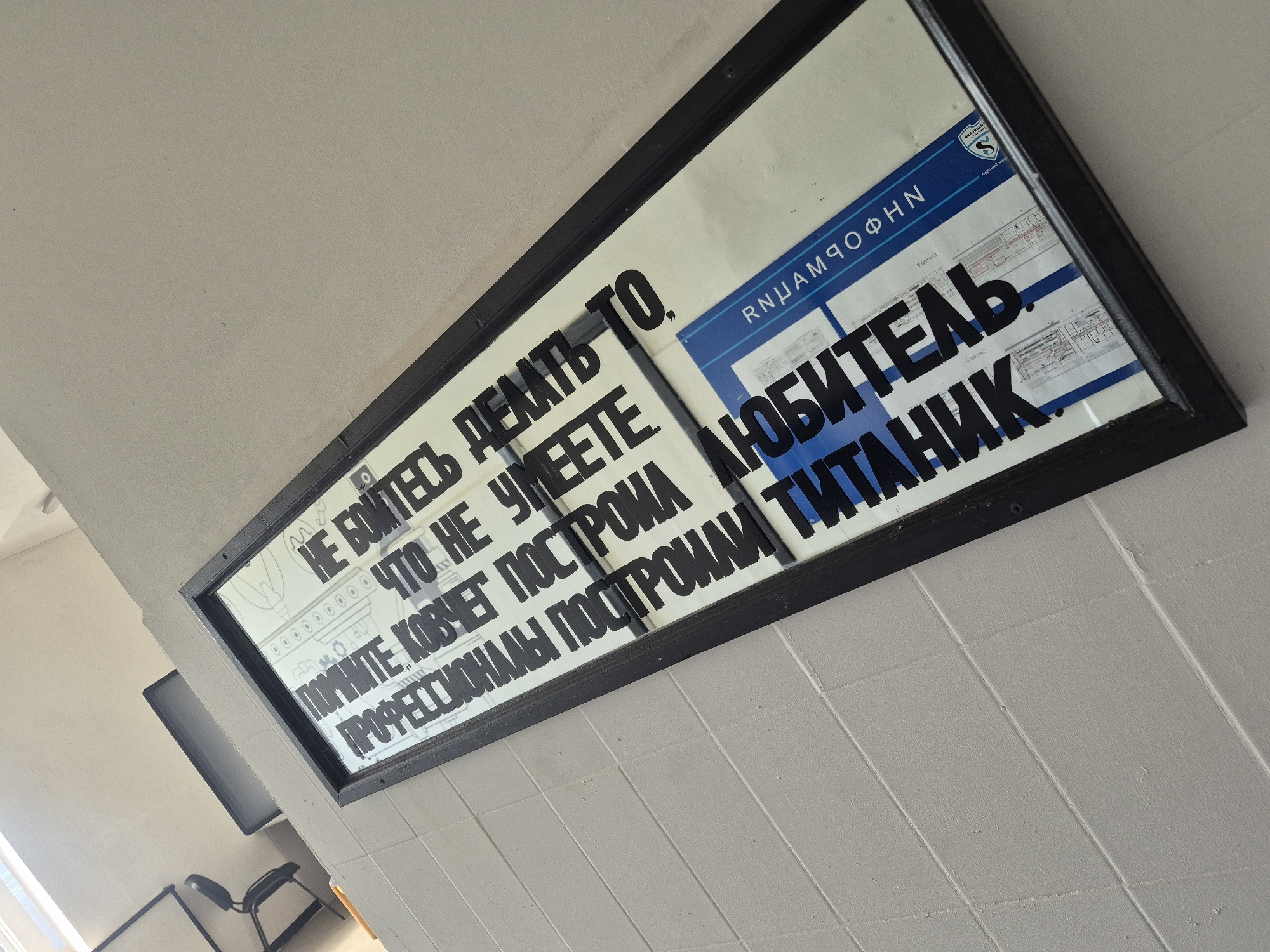 The image shows a black-framed poster hanging on a light-colored tiled wall. The poster has text in Russian written in bold, black letters. Above the text is a drawing of an ark. To the right of the text is a blue and white architectural drawing with the title "RNUAMQOFHN." A logo with the letter "S" is visible in the upper right corner. The text on the poster reads: "НЕ БОЙТЕСЬ ДЕЛАТЬ ТО ЧТО НЕ УМЕЕТЕ. ПОМНИТЕ КОВЧЕГ ПОСТРОИЛ ЛЮБИТЕЛЬ, ПРОФЕССИОНАЛЫ ПОСТРОИЛИ ТИТАНИК," which translates to "Don't be afraid to do what you don't know how to do. Remember, the Ark was built by an amateur, the Titanic was built by professionals."
The location is likely a public space such as a school or government building in Chaikovsky, Russia.