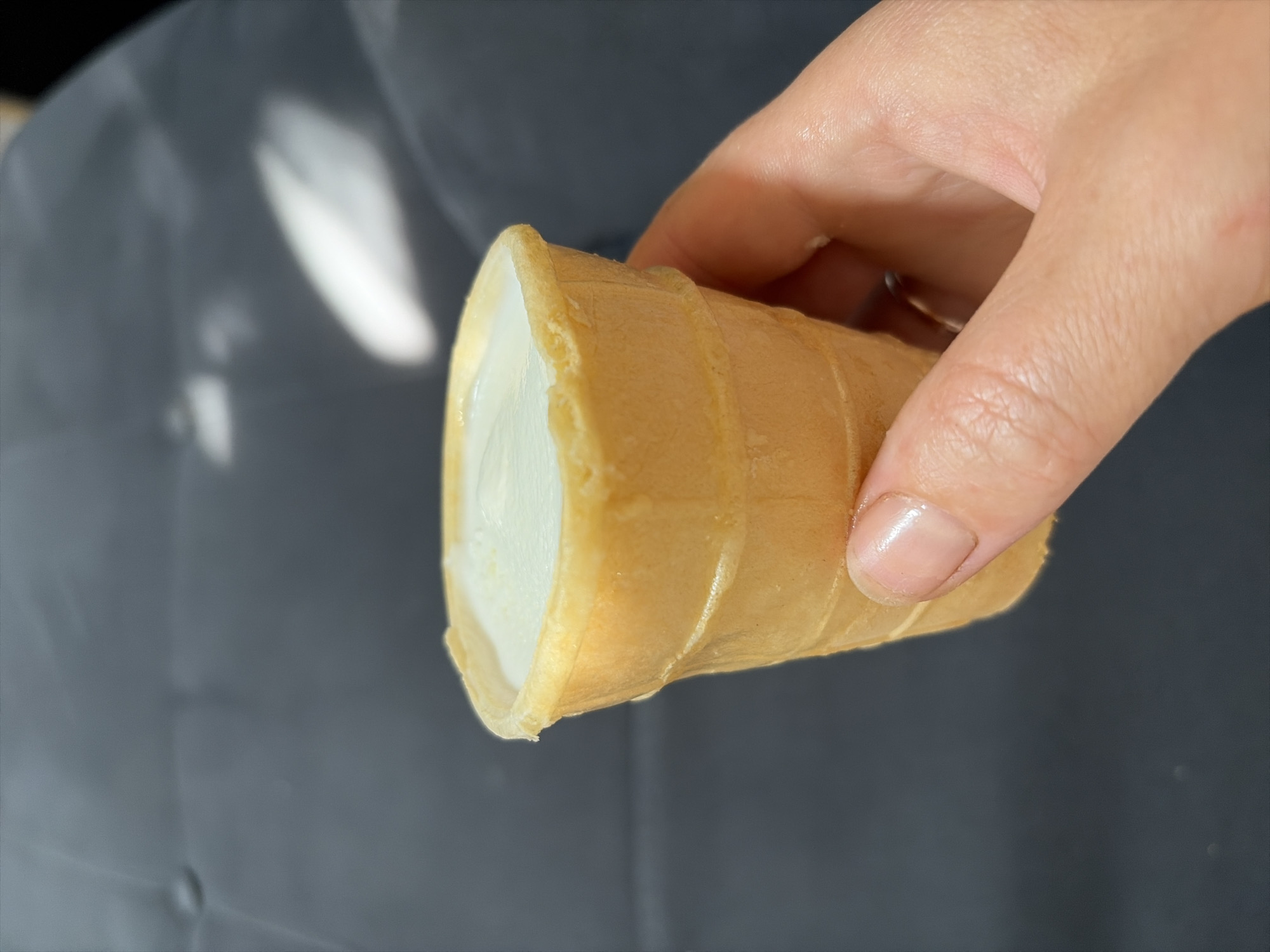 A person's hand is holding a mini ice cream cone filled with vanilla ice cream. The cone is made of a light brown waffle material and has horizontal ridges. The ice cream is white and has a smooth, creamy texture. The background is a dark gray, textured surface, possibly a sofa or chair. The image does not provide any information about the location in Oxentea, Moldova.