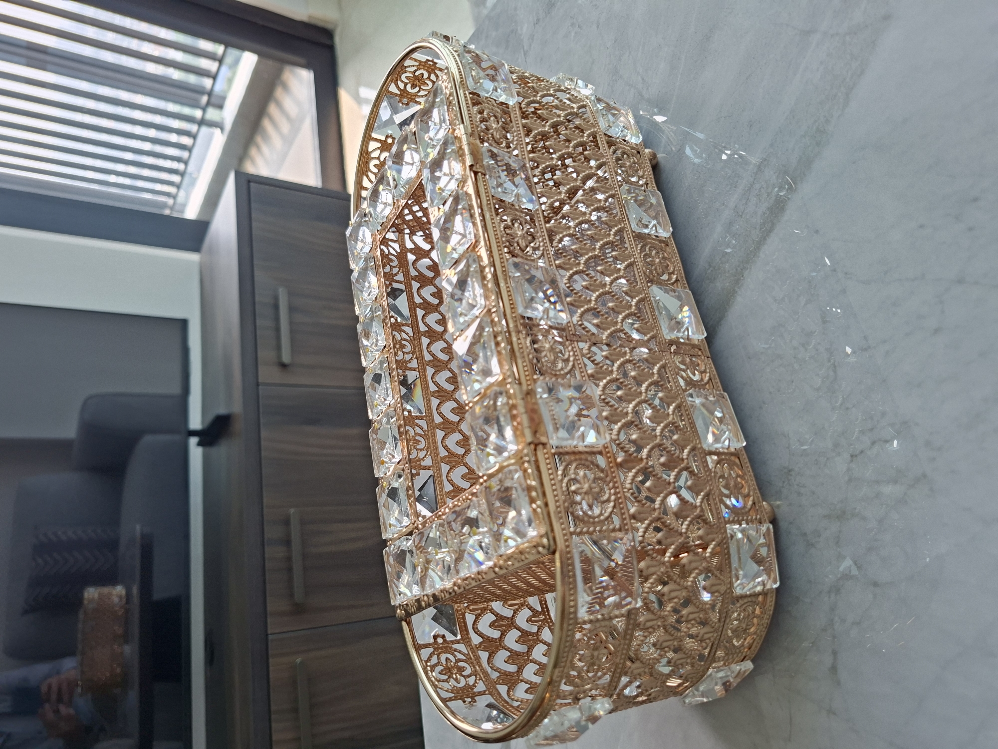 The image shows a crystal and gold decorative tissue box placed on a marble countertop. The tissue box is ornate, with intricate gold filigree and clear, square-shaped crystals embedded in its design. The background reveals a modern living room with a dark wooden TV console and a portion of a gray sofa. Sunlight streams in from a window with vertical blinds on the right, suggesting the location is likely a residential apartment. The image does not contain any information that can be used to determine its location in West Jakarta, Indonesia.