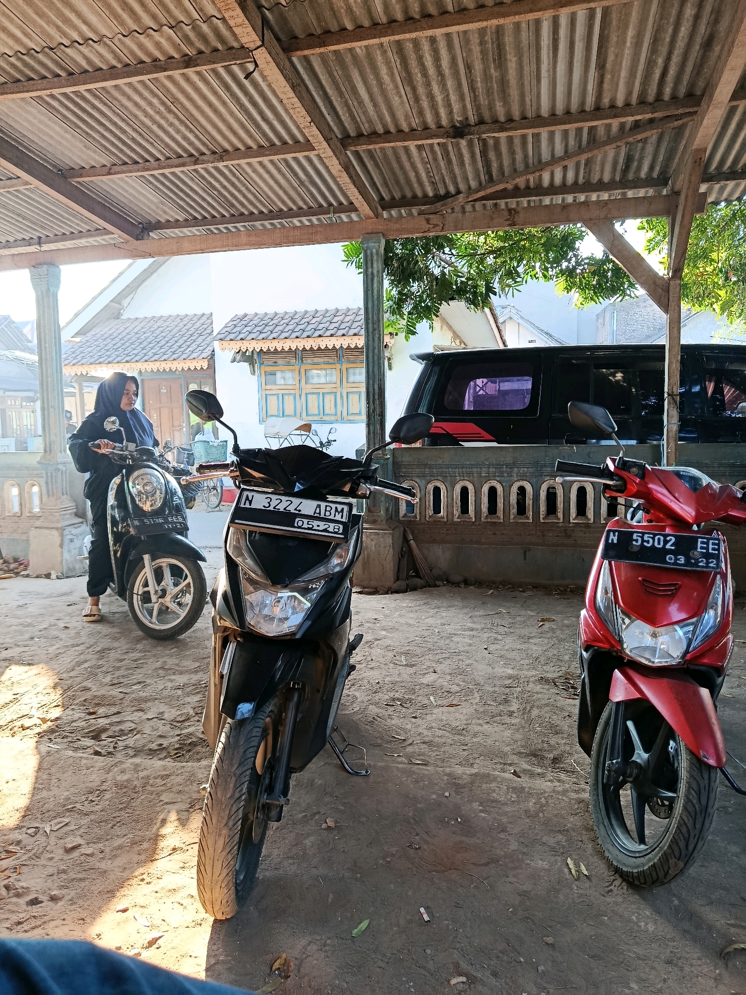 A woman wearing a hijab and dark clothing is standing next to a black scooter under a corrugated metal roof. The scooter has a license plate that reads "N 3222 ABM". In front of the black scooter is another scooter, a red one with a license plate reading "N 5502 EE". Behind the scooters, a black car is parked in front of a stone wall with arched openings. The ground is dirt and sand, and the scene appears to be in a rural area of Bululawang, Indonesia, evidenced by the architecture and the presence of scooters. The lighting suggests it is daytime, with sunlight casting shadows.