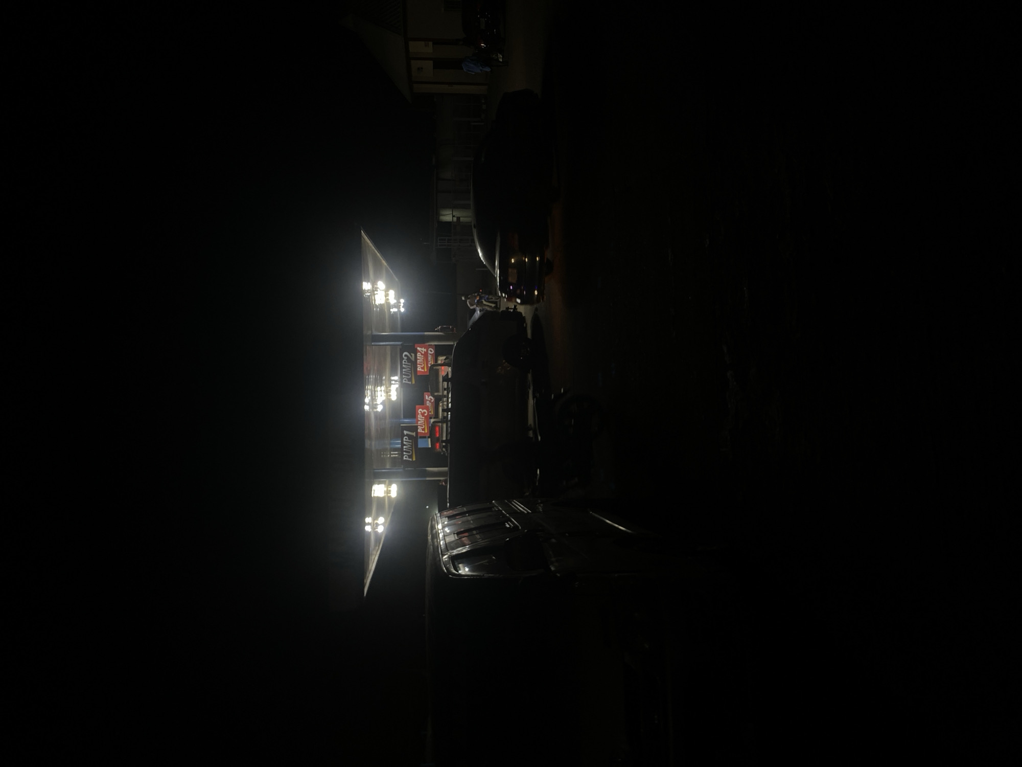 The image shows a gas station at night in Tudun Wada, Nigeria. The scene is dark, illuminated primarily by the bright lights of the gas station canopy. The canopy covers several gas pumps, labeled with numbers. A dark vehicle is parked near the pumps, and a person can be seen standing near the pumps as well. The surrounding environment is obscured by darkness.