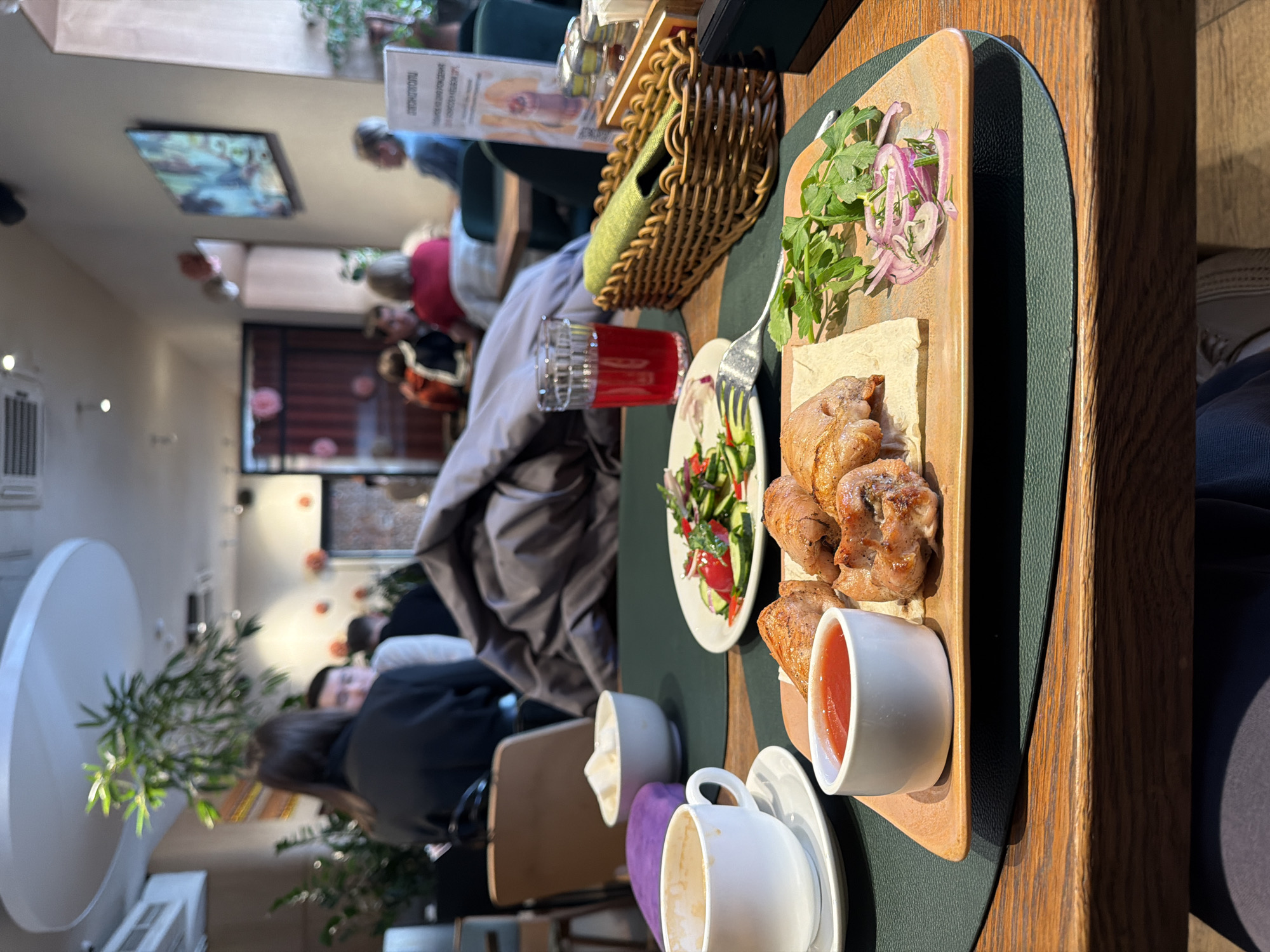 The image shows a table at a restaurant in Moscow, Russia. On the table is a wooden serving board with grilled meat, flatbread, red onion salad, herbs, tomato sauce, a small salad with tomatoes and cucumbers, a glass of red juice, and a basket, possibly with bread. There are also two white cups, one on a saucer.
Several people are sitting at nearby tables. There is a painting on the wall in the background and a plant in a white pot. The restaurant seems to be well-lit with a mix of natural and artificial light.