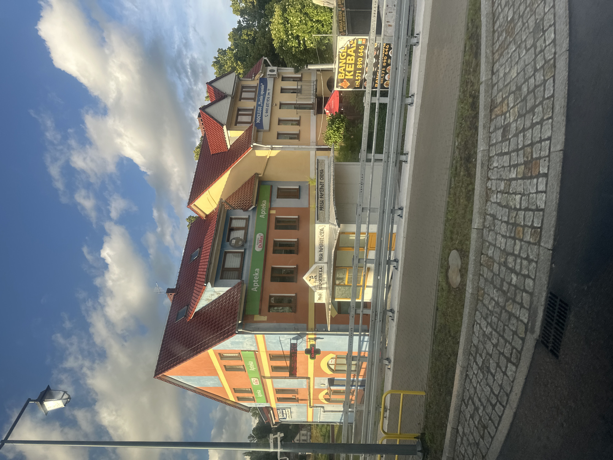 The image shows a colorful building complex in Krosno Odrzańskie, Poland. The building on the left is painted in vibrant orange and blue stripes with a red roof. It has several windows and signage for "Apteka" (pharmacy) and "HOSTEL". To the right of this building, there is another building painted in yellow with a red roof, featuring signage for "NOCLEGI <<Tanie spanie>>" (Accommodation <<Cheap sleep>>). A smaller structure in front of the buildings appears to be a kiosk or shop with signs that read "PRASA PAPIEROSY CHEMIA" (Press, cigarettes, chemistry) and "KOLPORTER SA". Further to the right, there is a sign for "BANGLA KEBAB" with a phone number. The buildings are situated along a road with a metal guardrail and a cobblestone sidewalk in the foreground. The sky above is blue with scattered white clouds, and trees are visible in the background. The overall scene depicts a commercial area with various businesses.
