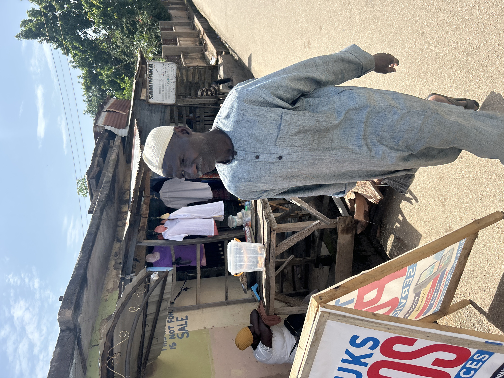 In Jos, Nigeria, an elderly man wearing a traditional light blue robe and a white cap walks along a paved road. He is seen from the chest up, looking to the left of the frame. Behind him, a small market stall features various clothing items, including white and purple shirts, hanging from a wooden structure. A sign on the stall reads "THIS IS NOT FOR SALE." To the left of the main subject, another person, wearing a yellow cap, is slumped over a table, appearing to rest. A wooden A-frame sign stands in the foreground, displaying advertisements in red and blue lettering. The scene unfolds under a partly cloudy sky, with lush green trees visible in the background, suggesting a warm, sunny day.