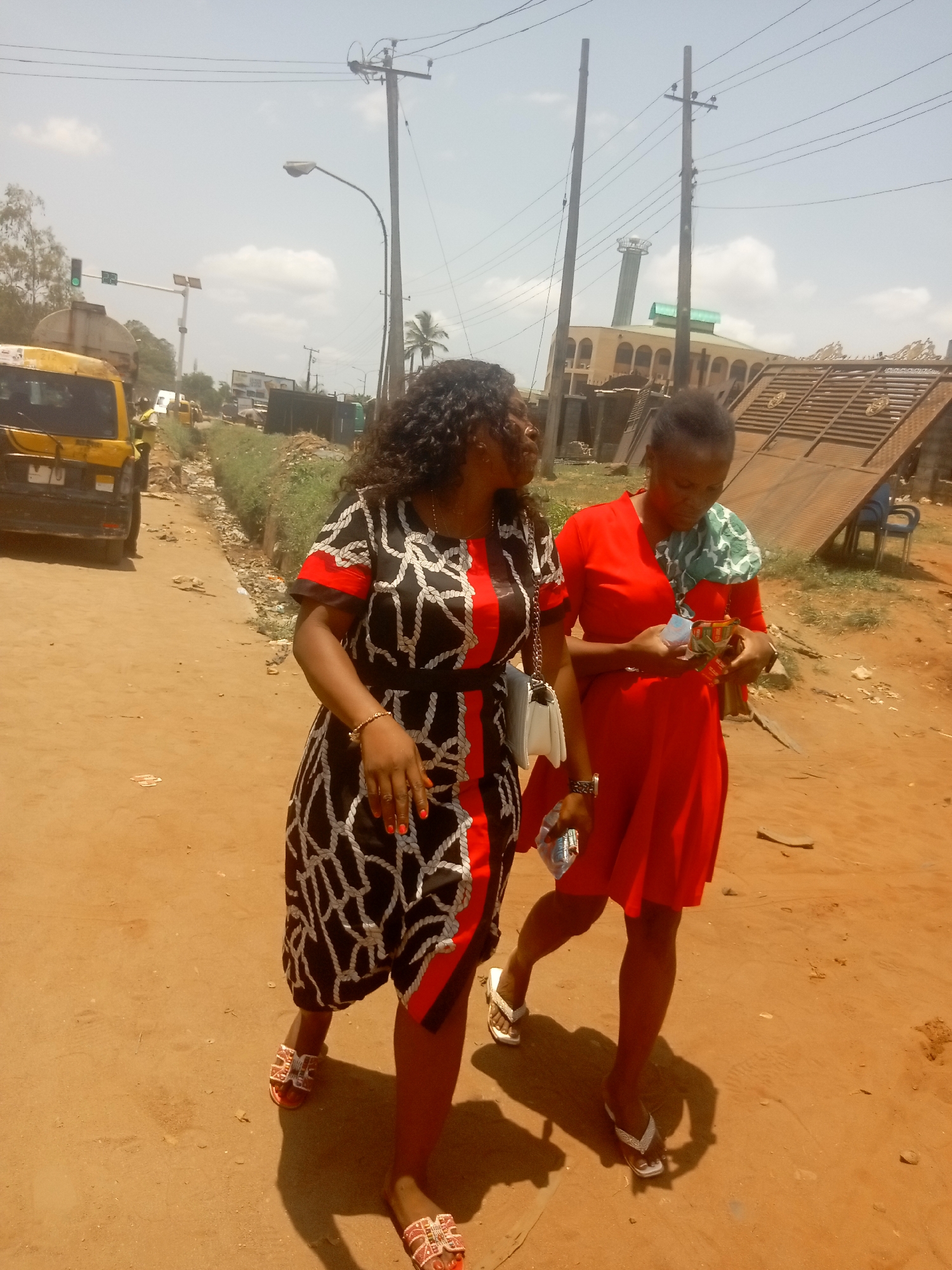 Two women walk along a sunny, unpaved dirt road in what appears to be an urban or semi-urban area, identified as Onitsha, Nigeria. The woman on the left has dark curly hair and wears a black dress adorned with white rope-like patterns and red stripes. She carries a white handbag, sports red nail polish, and wears patterned sandals. The woman on the right, with her dark hair tied back, is dressed in a plain red dress with a green and white patterned fabric draped over her shoulders. She wears white flip-flops and is focused on items held in her hands, possibly engaging in conversation with the other woman.
The scene is brightly lit by the sun, casting strong shadows, indicating midday or early afternoon. The sky is clear with scattered clouds. In the background, a yellow commercial van or bus is visible on a more defined road. Utility poles with a dense network of power lines crisscross the sky, alongside a street lamp. Further down the road, there's a prominent building featuring a tall minaret, suggesting a mosque. Adjacent to it, a metallic gate leans against another structure, near which two blue plastic chairs are placed. An overgrown ditch runs alongside the dirt path. Visible text "MO 70" can be seen on the front bumper of the yellow van. Other distant text on signs and on the items held by the woman in red are illegible. The overall atmosphere suggests a casual, everyday street scene.
