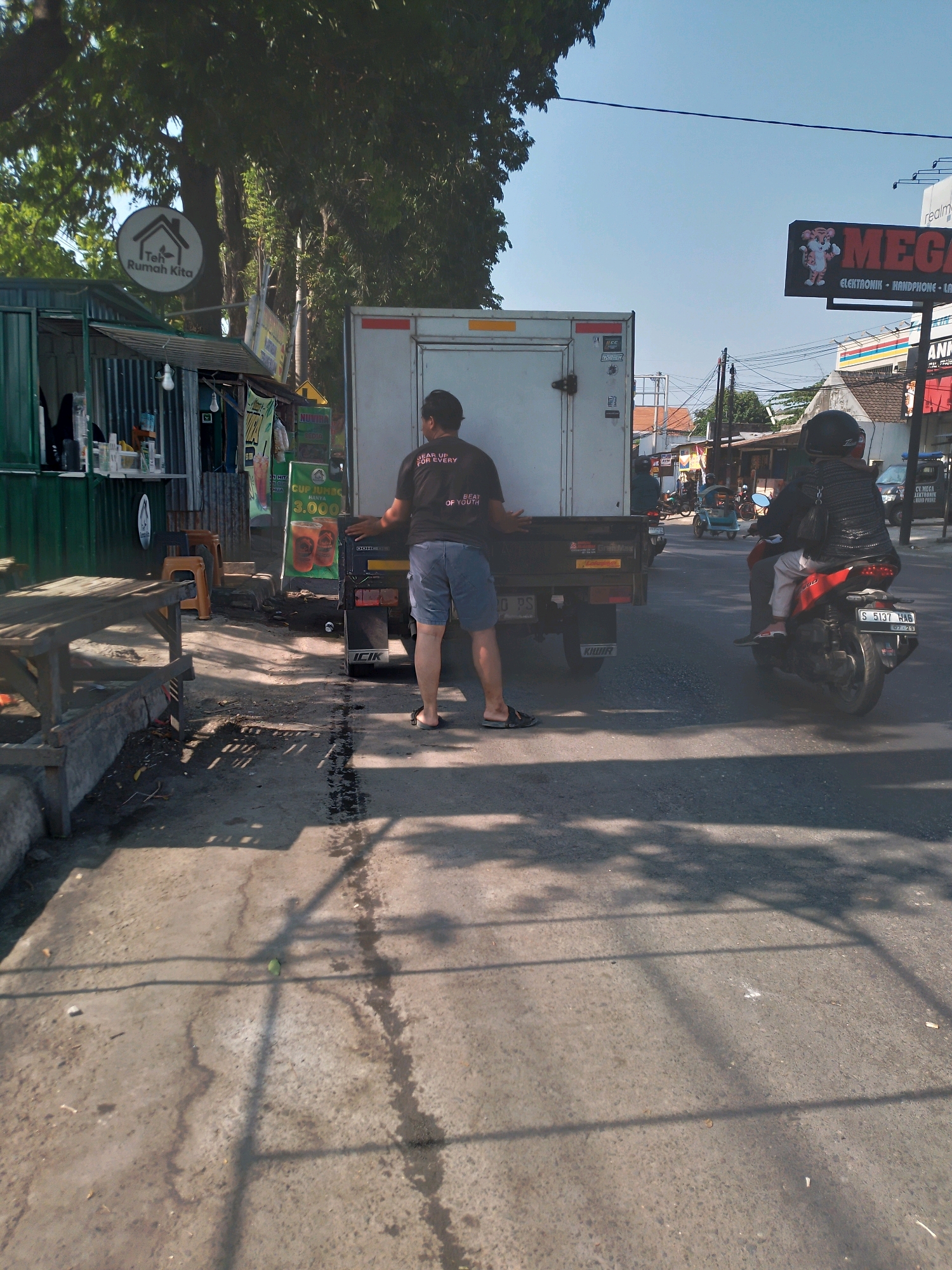 The image depicts a street scene in Kertosono, Indonesia, on a sunny day. In the foreground, a man wearing a black t-shirt and blue shorts is standing next to the back of a white truck with a closed cargo container. The truck has the word "KIJAR" visible on its rear. To the left of the truck, there is a green roadside stall with tables and chairs. A sign with a house and the text "Teh Rumah Kita" is visible above the stall. Further down the street, a motorcycle with two occupants, a driver wearing a helmet and a passenger, is moving away from the camera. The passenger is wearing a striped jacket. A blue three-wheeled vehicle is also visible in the distance. The street is paved and shows signs of wear, with some water or oil stains. Shadows from trees and possibly overhead wires are cast across the scene. In the background, there are buildings, trees, and utility poles, typical of a town or city environment. A large billboard advertising "MEGA" electronics is visible in the upper right portion of the image. The sky is clear and blue.