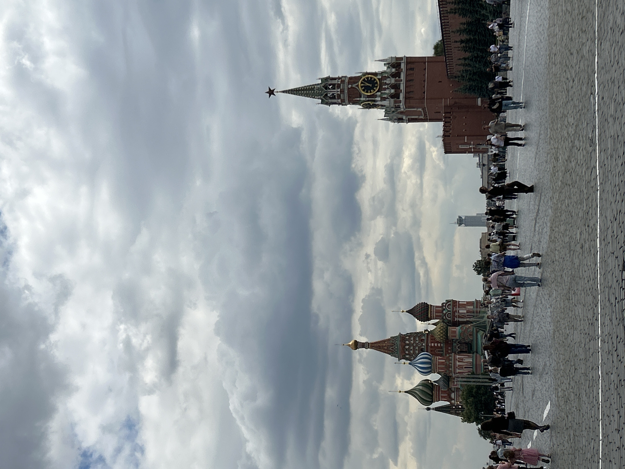 The image shows a wide view of Red Square in Moscow, Russia, under a cloudy sky. In the foreground, cobblestones pave a large open area with a white line marking a section of the square. Numerous people are scattered throughout the square, walking in various directions.
To the left, the iconic St. Basil's Cathedral is visible with its distinctive multi-colored onion domes. To the right, the Spasskaya Tower, a prominent clock tower of the Moscow Kremlin, stands tall with its red brick facade and a red star at its apex. Behind the Spasskaya Tower, a segment of the Kremlin wall with evergreen trees in front of it is visible. In the background, a modern tall building can be seen rising above the cityscape.