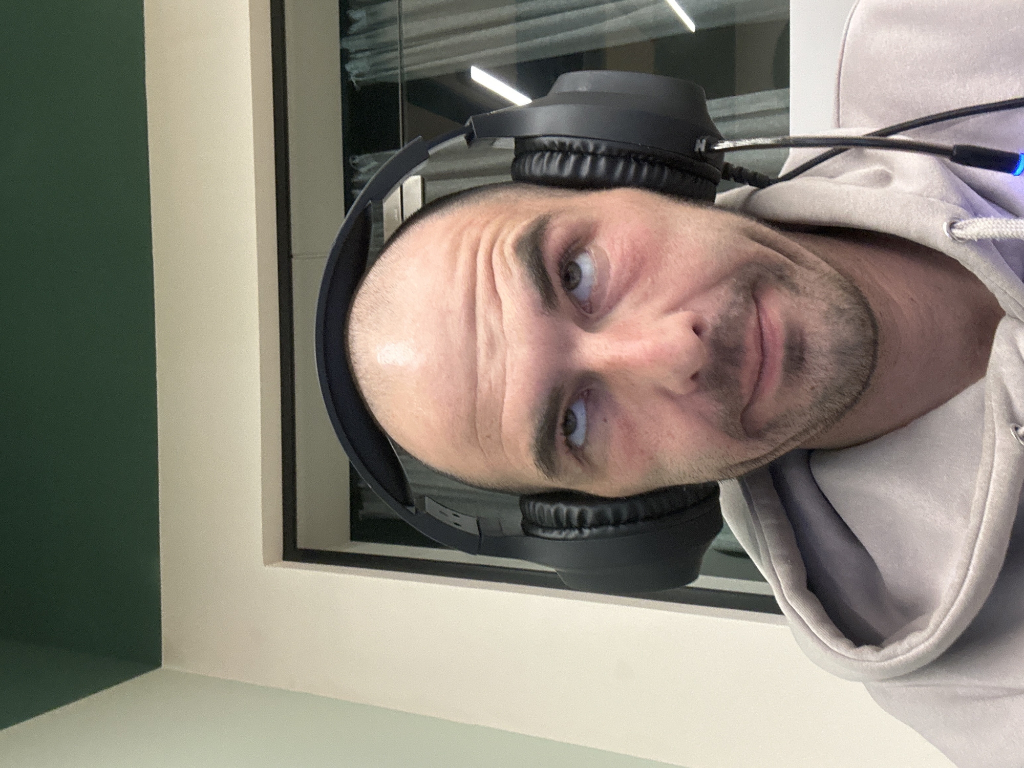 A close-up portrait shows a bald man wearing black headphones and a gray hoodie. He appears to be indoors near a window. The image does not show any information that would identify the location as Budva, Montenegro.