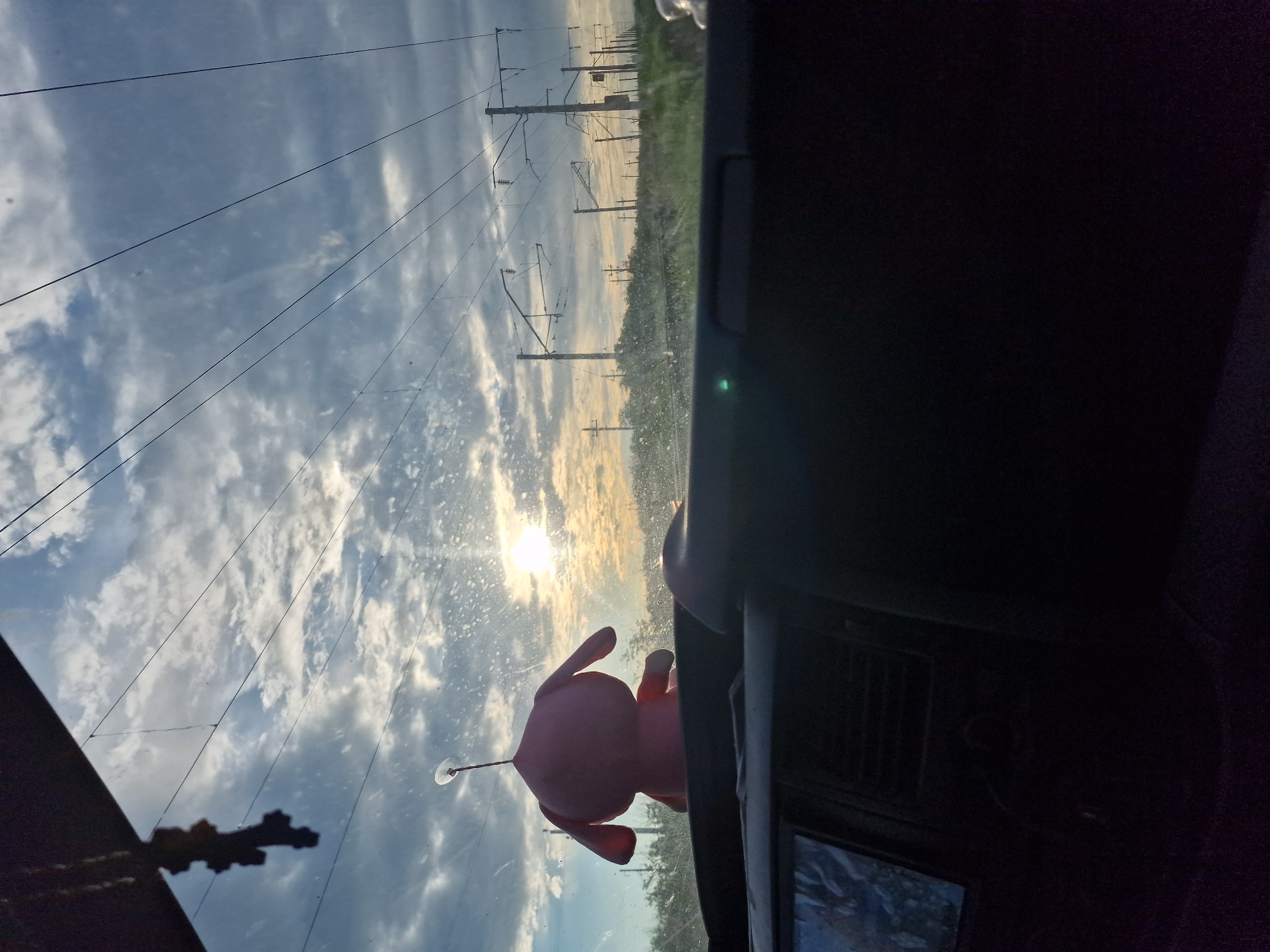 The image, taken from inside a vehicle looking out the windshield, depicts a scenic view of the sky over Kursavka, Russia. The sky is a dramatic mix of clouds and sunlight, with rays of light breaking through. Overhead, power lines stretch across the frame, supported by utility poles visible in the distance. In the foreground, a pink plush toy is attached to the windshield by a suction cup. The lower portion of the image shows the interior of the vehicle, including the dashboard and a screen. The overall impression is one of a journey through a landscape marked by both natural beauty and infrastructure.