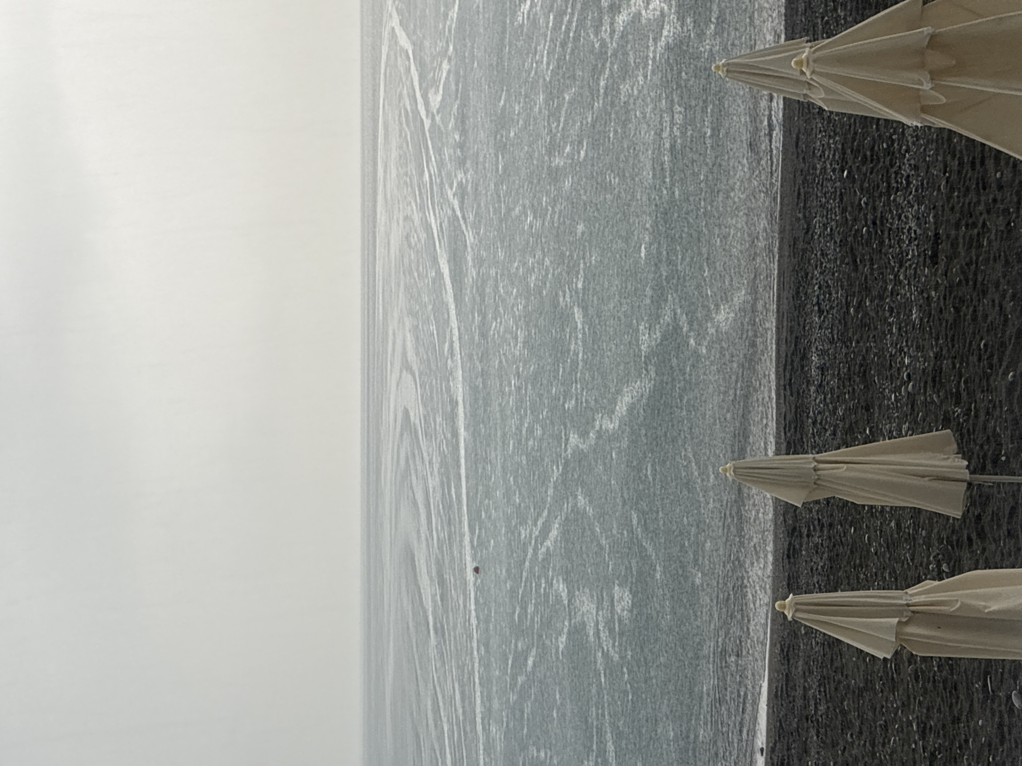 The image depicts a serene, overcast scene at a beach, likely in Sochi, Russia, given the context. In the foreground, three closed beach umbrellas stand on a dark, pebbly shore. The umbrellas are cream-colored and their fabric is gathered, obscuring their full form. Beyond the beach, the sea stretches out, its surface marked by subtle, swirling patterns, suggesting a gentle current or wind. The water appears choppy and dark gray under the diffused light of the sky. A small, red object, possibly a buoy or a distant boat, is visible in the middle ground of the sea. The horizon line is indistinct due to the overcast conditions, blending the sea and sky into a uniform expanse of muted gray. The overall impression is one of quietude and a cool, perhaps slightly damp, atmosphere.