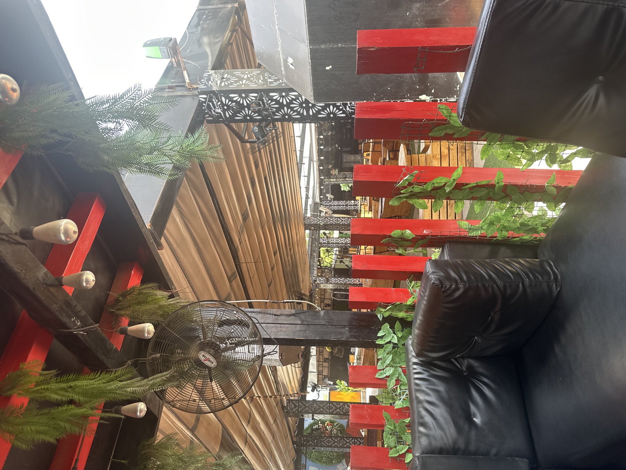 The image depicts an outdoor seating area, likely a restaurant or bar, with a rustic yet modern aesthetic. The primary subjects are black leather armchairs, some with visible stitching and tufted details, positioned on a deck. Red painted wooden beams and slats form decorative and structural elements throughout the scene, interspersed with artificial green foliage, creating a vibrant contrast. A large, circular industrial fan is mounted on a black beam, suggesting a warm climate.
The scene is oriented vertically, with the view looking upwards and downwards along a pathway or seating arrangement. Above, a light-colored awning stretches across, and hints of a bright, overcast sky are visible. Below, more red beams and wooden structures can be seen, along with a glimpse of more seating and decorative elements. A black metal railing with an intricate floral cutout pattern adds a touch of elegance.
There are no people visible in the image, and no specific activity or event is taking place. The lighting suggests daytime, possibly late morning or afternoon, with diffused light indicating an overcast day. The presence of the fan and abundant foliage points towards a tropical or subtropical location. The text "KENSTAR" is visible on the fan. The location context provided, "Howayi, Nigeria," aligns with these environmental cues. The overall impression is a stylish, comfortable outdoor space designed for relaxation.