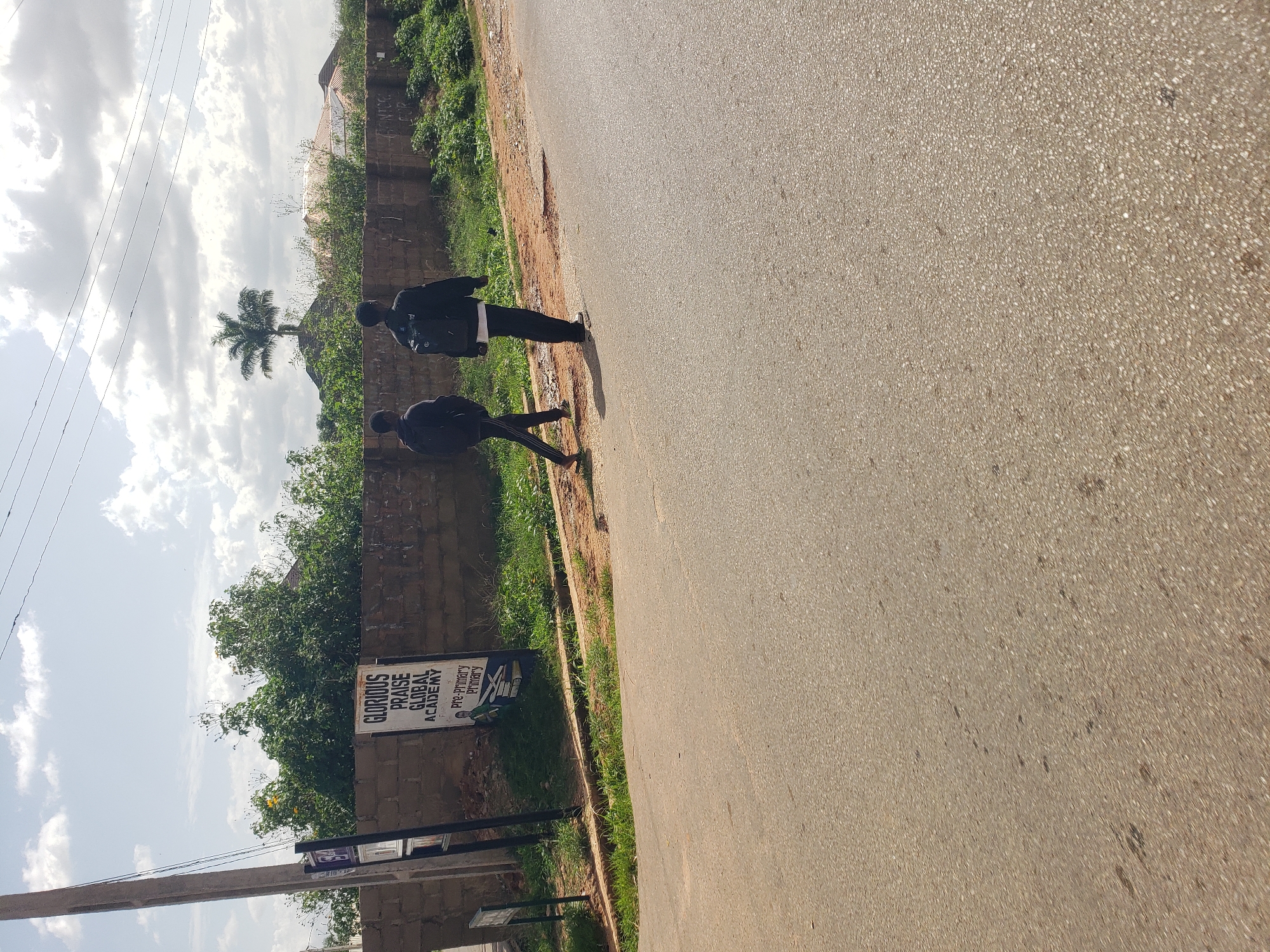 Two young males, dressed in casual clothing, are walking away from the camera on a paved road. The male on the left is wearing a dark hoodie and black pants with a white stripe down the side. The male on the right is wearing a black jacket over a white t-shirt and black pants. They are walking on the right side of the road.
To the left of the road, there is a brown brick wall bordering a grassy area. A sign with text "GLORIOUS PRAISE GLOBAL ACADEMY" is visible on the wall, along with an image of a student reading books. A utility pole is situated to the left of the sign.
Beyond the wall, lush green trees and vegetation are abundant. In the background, several buildings with light-colored roofs are visible, and a single palm tree stands tall against a cloudy sky. The scene appears to be a street in Gura Top, Nigeria. The overall lighting suggests it is daytime.