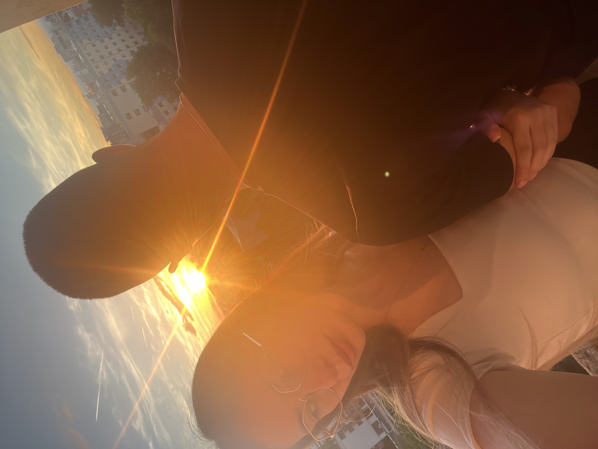 A low-angle, full-frame, outdoor, close-up shot captures two people against a sunset sky. The person on the right is a male with a shaved head, seen from the back. He is wearing a black t-shirt and a thin silver chain necklace. The person on the left is a female, facing forward and wearing eyeglasses with gold frames, and a white top. Her hair is light brown and long. Her left arm is resting on a railing or ledge, and her right hand is held by the male. The sunset is a bright orange and yellow, with clouds and light rays creating a lens flare effect that obscures parts of the image. In the background, a city skyline with buildings and trees is visible. The lighting suggests it is late afternoon or early evening.