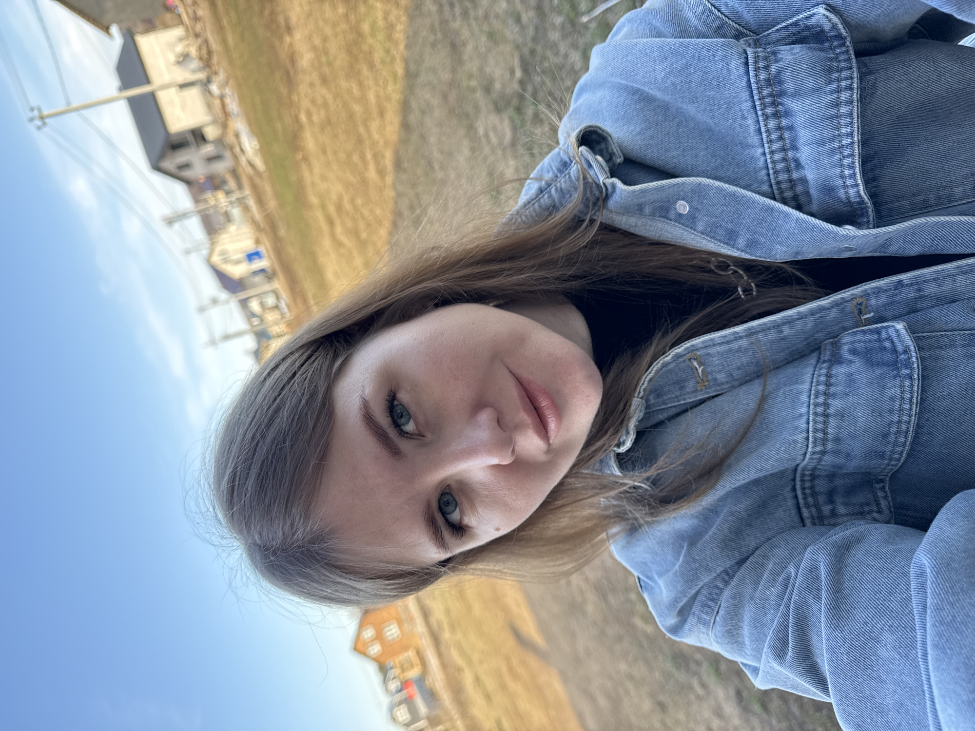 A woman with long brown hair is wearing a light-wash denim jacket. She is outdoors, likely in a rural or suburban area of Gornyi (Dvurechenskoe S/P), Russia. In the background are houses and utility poles; there is a field or open space. The sky is light blue. The photo is taken from a low angle.