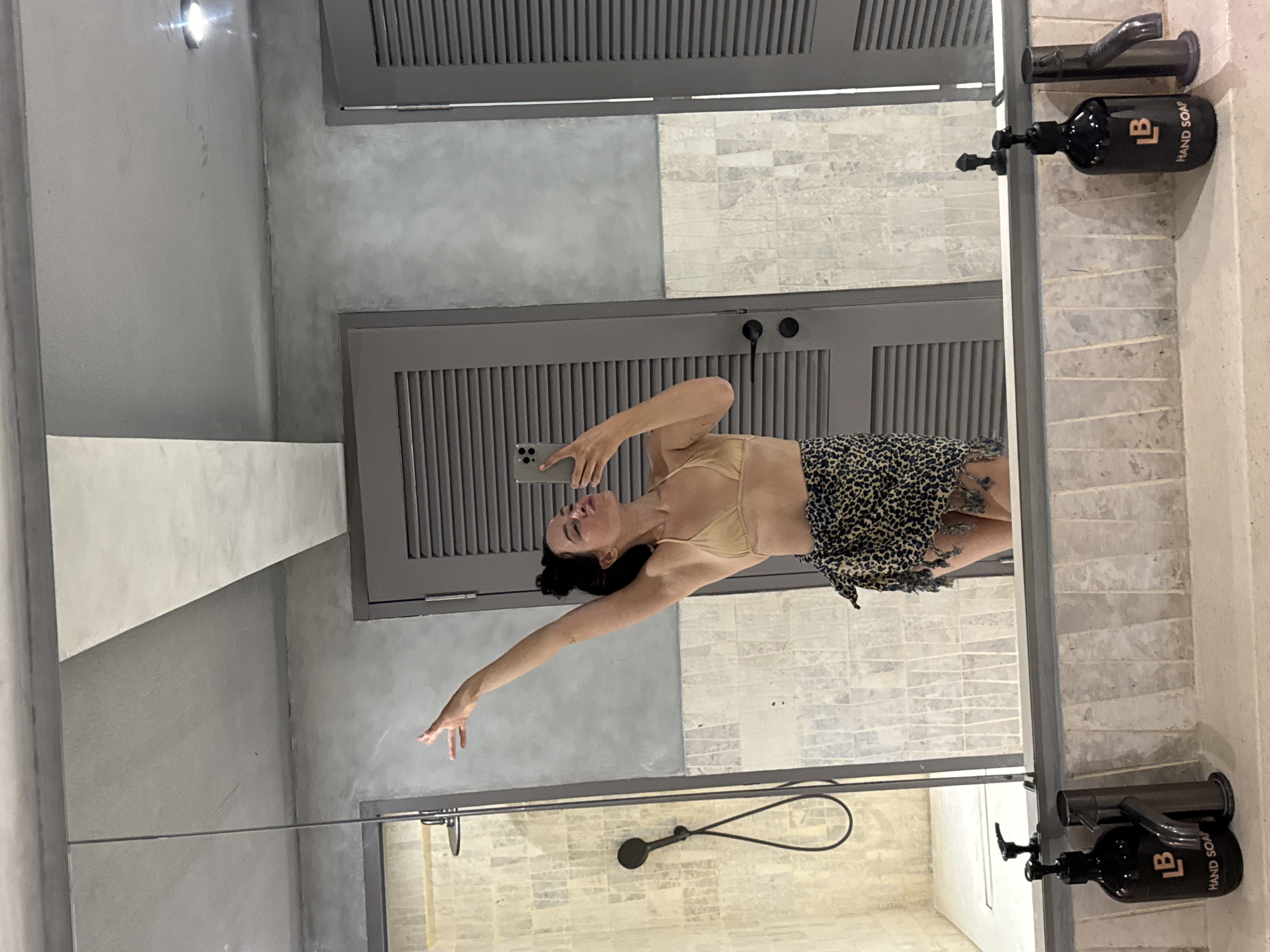 A young woman with dark hair in a bun takes a mirror selfie in a modern bathroom. She is wearing a tan bikini top and a leopard print sarong. Her right arm is raised with her hand in the air, and her left hand holds a smartphone to take the picture. She is pouting her lips. The bathroom features a tiled shower with a black handheld showerhead, grey louvred doors, and a grey concrete ceiling. A large mirror above a countertop reflects the scene. Two black soap dispensers with the logo "LB HAND SOAP" and two faucets are visible on the countertop in the foreground. The overall aesthetic is minimalist and contemporary.