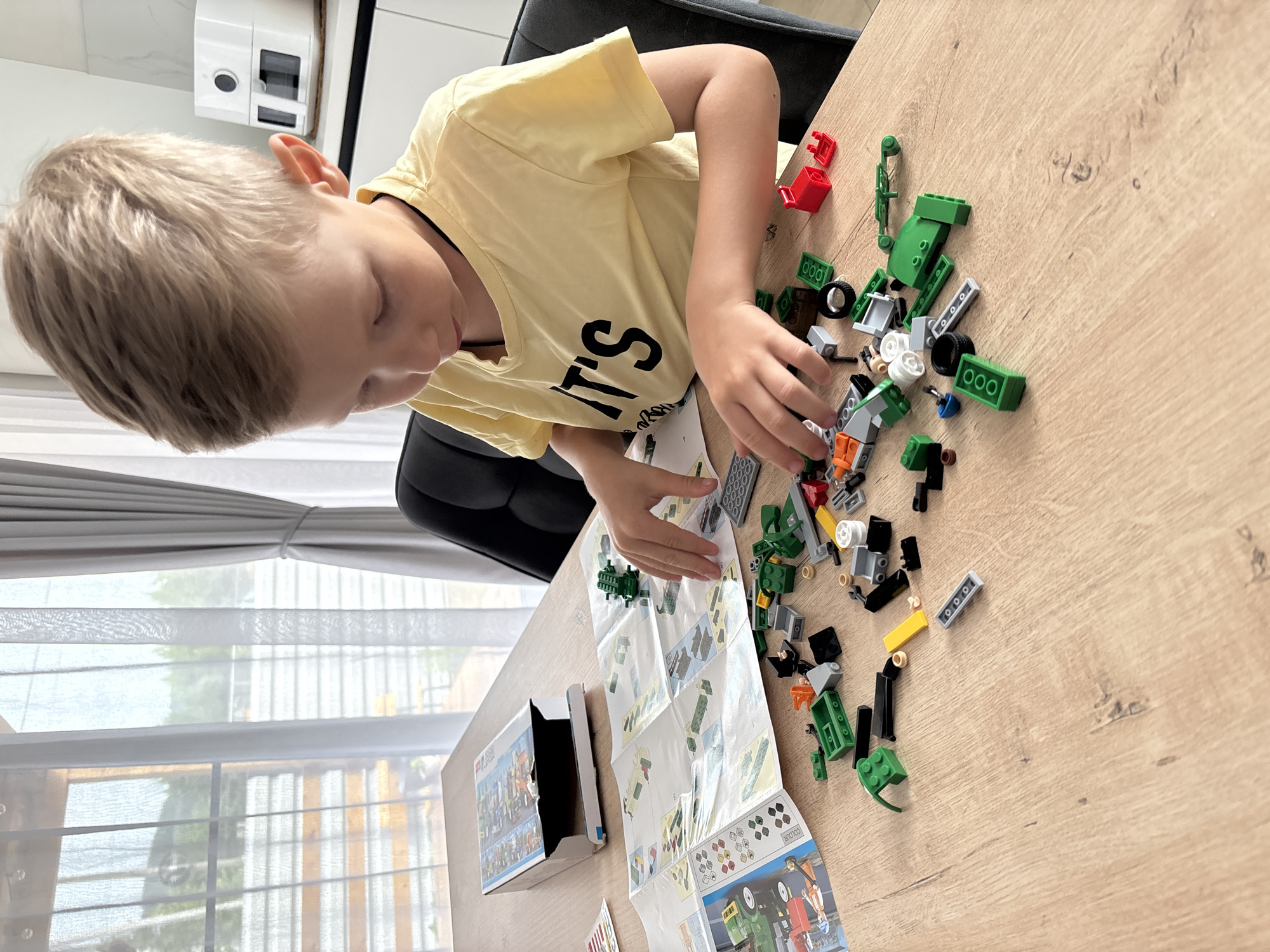 A young boy is engrossed in building with LEGOs on a wooden table. He is wearing a yellow t-shirt with "IT'S" printed on it in black. The table is covered with various LEGO bricks, primarily in green and red, along with instruction manuals. The scene appears to be indoors, with a window showing a blurred view of trees and buildings outside, suggesting it is daytime. The boy's focused expression and the scattered building blocks indicate he is actively engaged in play. A portion of a white box with LEGO branding is visible on the left side of the image. The overall setting seems to be a home environment.
