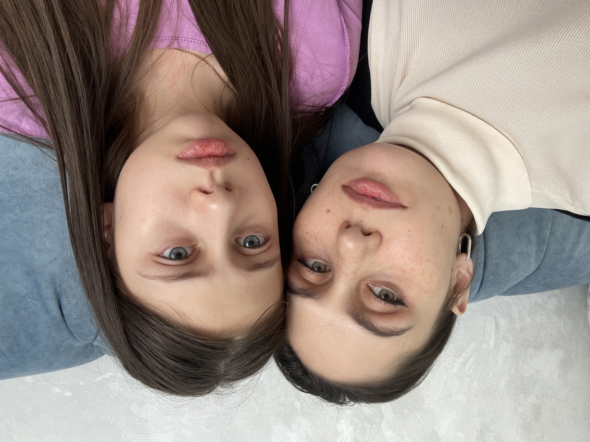 The image displays a close-up portrait of two individuals, a woman and a girl, in an indoor setting within the city of Izhevsk, Russia. Both subjects are positioned facing directly towards the camera, with their heads close together.
The individual on the left is an adult woman with dark brown hair, green eyes accented with black eyeliner, and a light skin tone featuring visible freckles across her cheeks and nose. She wears a cream-colored ribbed turtleneck garment, with a dark fabric visible beneath or to her left, and a silver-colored, oval-shaped hoop earring in her visible left ear. Her lips are slightly parted, and her expression is neutral to slightly serious.
The individual on the right is a girl with long, straight dark brown hair and blue eyes. She wears a lavender or light purple long-sleeved top and a thin gold-colored chain necklace around her neck. Her mouth is slightly downturned, forming a subtle pout, indicating a serious or somber expression.
The background consists of a textured light gray wall behind a segment of blue upholstered furniture, possibly a sofa or chair back. The lighting is uniform and diffused, illuminating the subjects evenly.