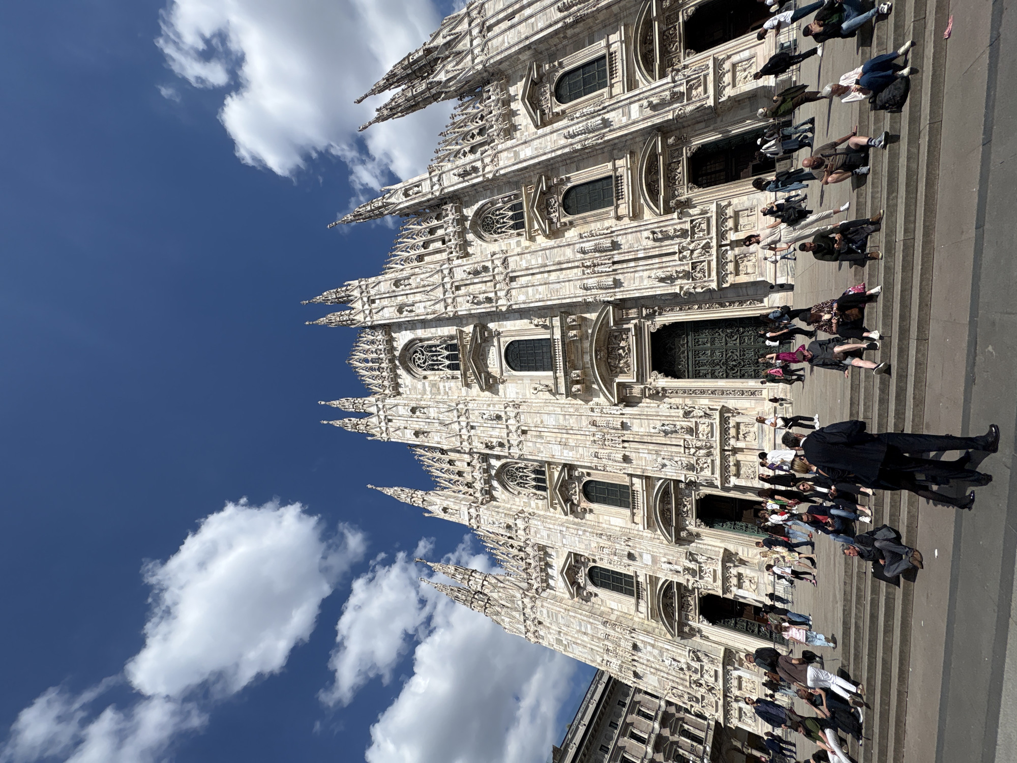 The image shows the Duomo di Milano, a large Gothic cathedral in Milan, Italy. The cathedral's ornate facade, constructed of white marble, dominates the frame, extending towards a blue sky dotted with fluffy white clouds. At the base of the cathedral, a large set of stone steps lead up to the entrance. Many people are sitting, standing, and walking on the steps, engaged in various activities, such as taking photos and conversing. One person wears a dark suit and stands in the foreground. A building is also visible in the lower left corner.