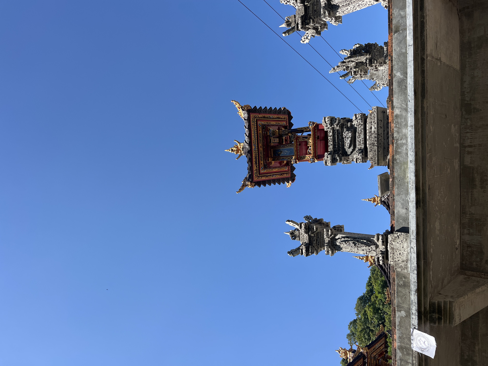 The image shows a clear blue sky above a rooftop in Karangasem, Indonesia. The rooftop features several intricately carved stone pillars, some topped with decorative elements in red and gold. The central structure is a multi-tiered pagoda-like building with ornate gold detailing. In the distance, lush green trees are visible, suggesting a tropical environment. Power lines run horizontally across the sky. A small white flag with an emblem is attached to the edge of the rooftop structure in the foreground. There are no people visible in the image. The scene appears to be a religious or cultural site.