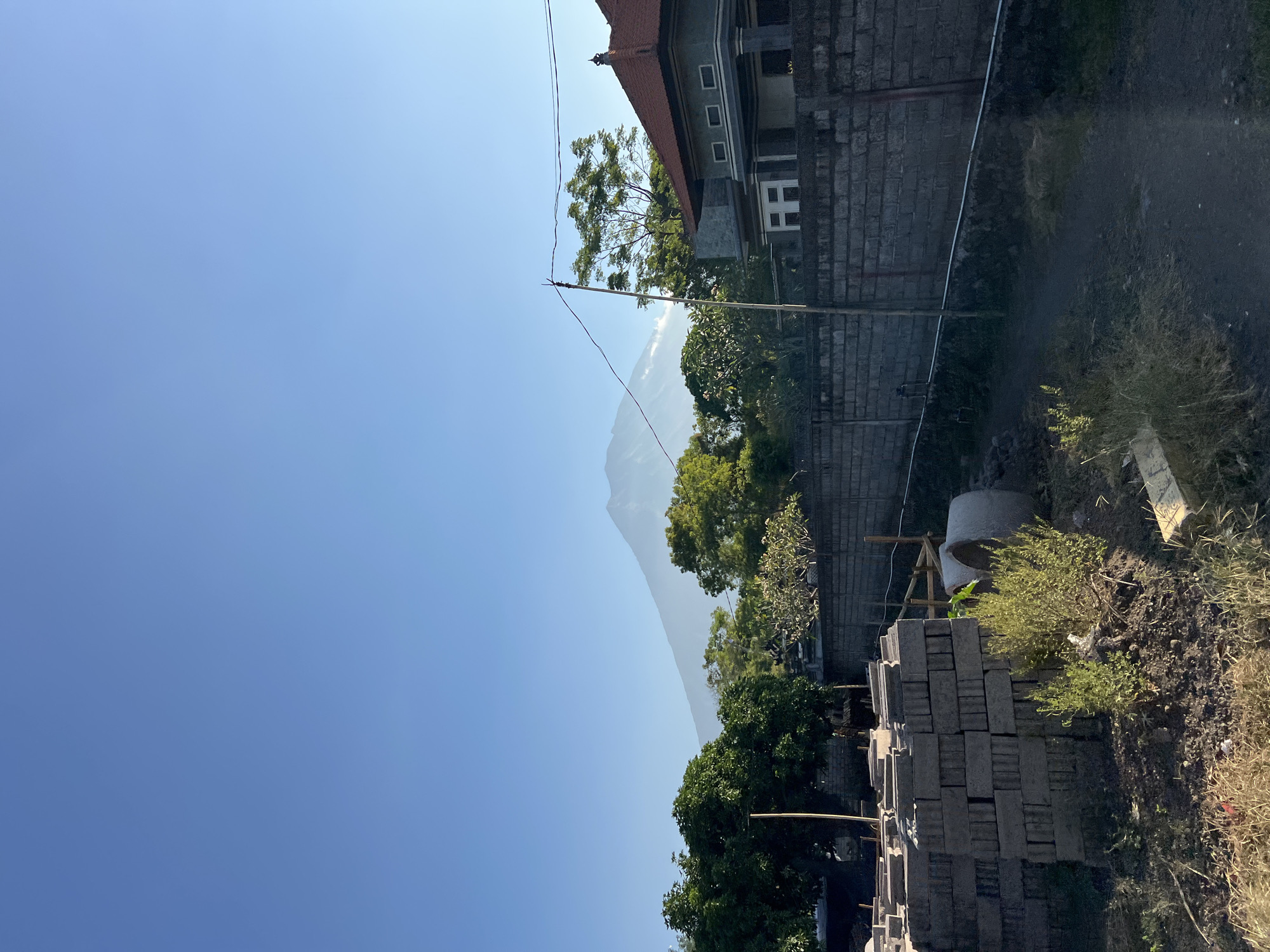 The image depicts an outdoor scene in Kubu, Indonesia. The foreground on the right shows a pile of gray bricks, some dry grass, and disturbed earth, suggesting a construction or renovation site. Behind the bricks, there is a large concrete pipe, possibly for drainage or construction. Further into the scene, a low brick wall separates the construction area from a building with a red tiled roof. This building appears to be made of dark stone or brick and has several windows. Beyond the building, lush green trees are visible, with a majestic mountain peak rising in the distance, partially obscured by haze. The sky is a clear, bright blue, indicating a sunny day, likely in the late morning or early afternoon given the sunlight's angle. There are power lines running across the scene, typical of a semi-urban or rural setting. The overall impression is one of ongoing construction set against a backdrop of natural beauty, characteristic of a tropical or subtropical location like Indonesia. There is no text visible in the image.