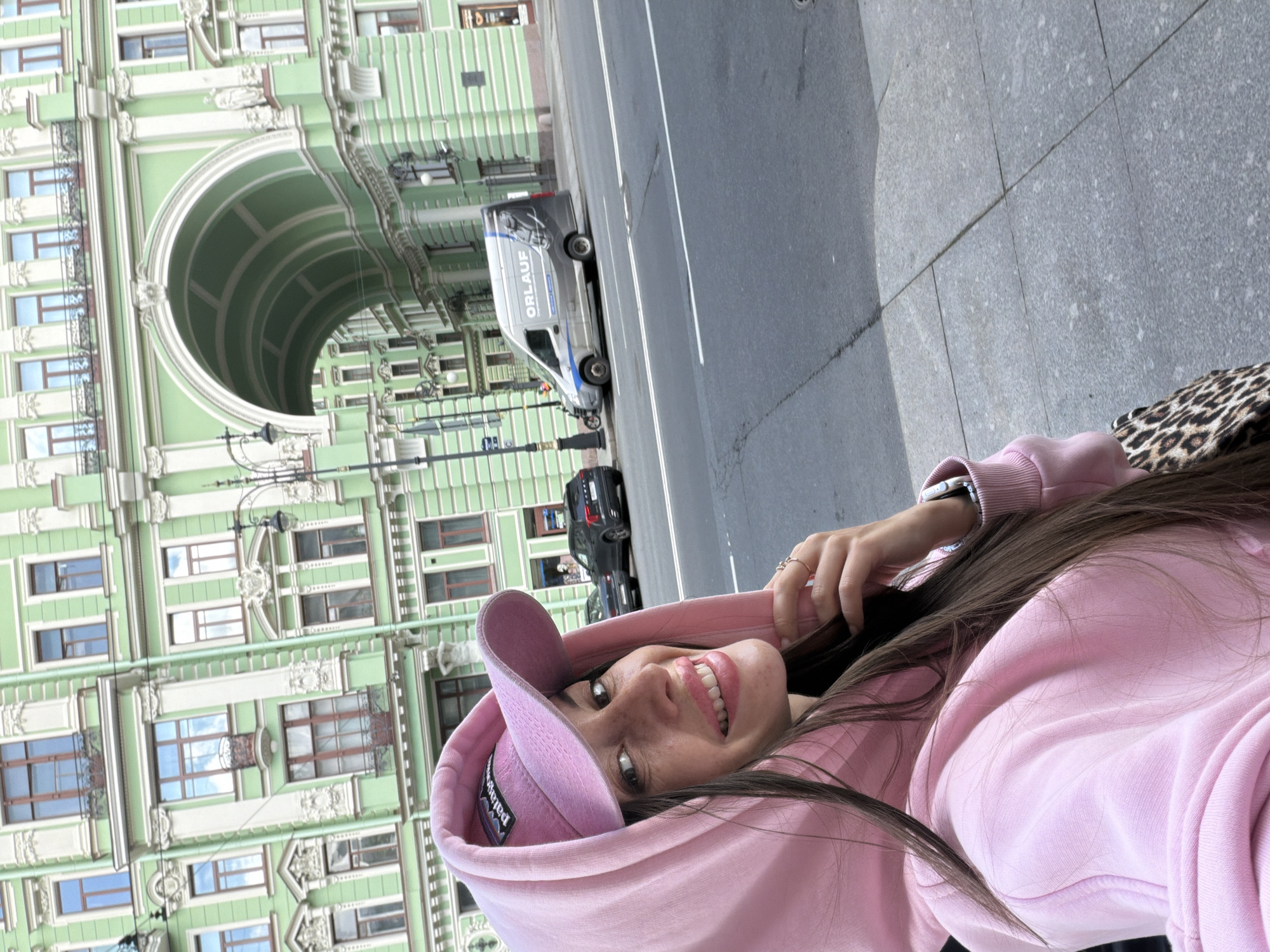 A woman is taking a selfie in front of a green arched building. She is wearing a pink hoodie and a pink baseball cap. She has long brown hair and is smiling. The building behind her is ornate and has many windows. There is a street in front of the building with cars and a sidewalk. The image appears to have been taken in Saint Petersburg, Russia. The building is likely the Singer House or one of its adjacent structures, recognizable by its distinct green facade and the archway.