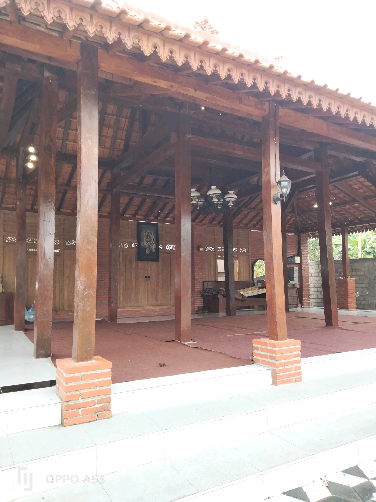 The image depicts the exterior of a traditional Indonesian mosque or prayer hall, likely located in the city of Trenggalek, Indonesia. The structure features a prominent wooden roof with intricate carvings along the eaves, supported by thick, reddish-brown wooden pillars. The walls are constructed from brick, and large wooden doors are visible in the background. The floor is covered with a dark red carpet, suggesting it is an indoor space. Several vintage-style lamps are suspended from the ceiling, and a framed artwork depicting a tiger hangs on one of the walls. In the foreground, white tiled steps lead up to the structure, with some of the wooden pillars resting on brick bases. The overall impression is one of traditional Javanese architecture.