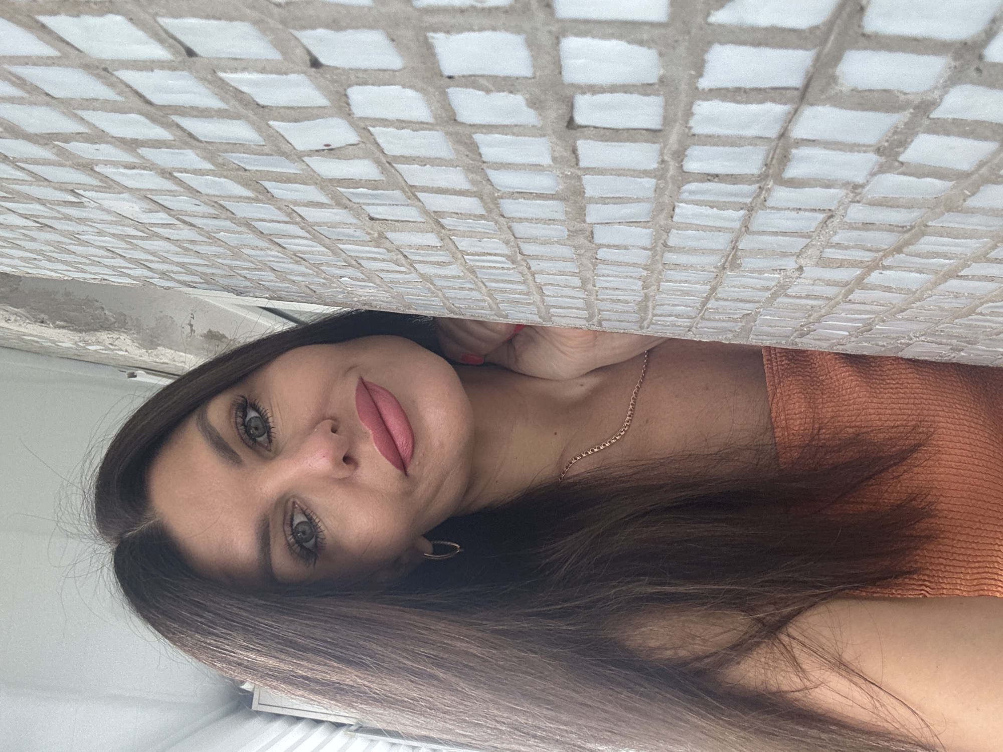 A woman with long, dark brown hair and light green eyes is leaning against a tiled wall. She is wearing an orange top and a gold chain necklace. Her right hand is touching her face. The background shows a window and what appears to be an apartment balcony. The location is identified as Chaikovsky, Russia.