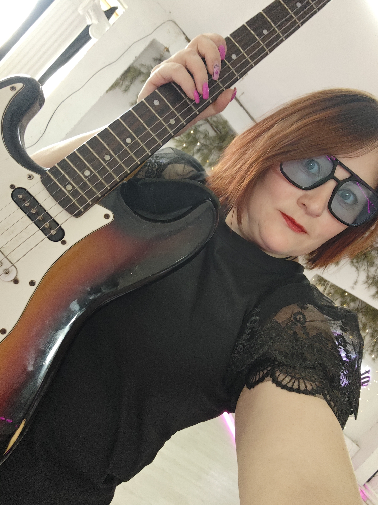 An individual with reddish-brown hair, blue eyes, and red lipstick is depicted indoors in Chaikovsky, Russia. The individual is wearing a black top with lace sleeves and black-framed glasses with blue-tinted lenses. Their left hand, adorned with pink nail polish, is positioned on the fretboard of a sunburst-finish electric guitar. The guitar features a white pickguard and three visible single-coil pickups. The background includes light-colored walls, green foliage decorated with string lights, and bright ambient lighting, indicating an indoor, possibly festive, setting.