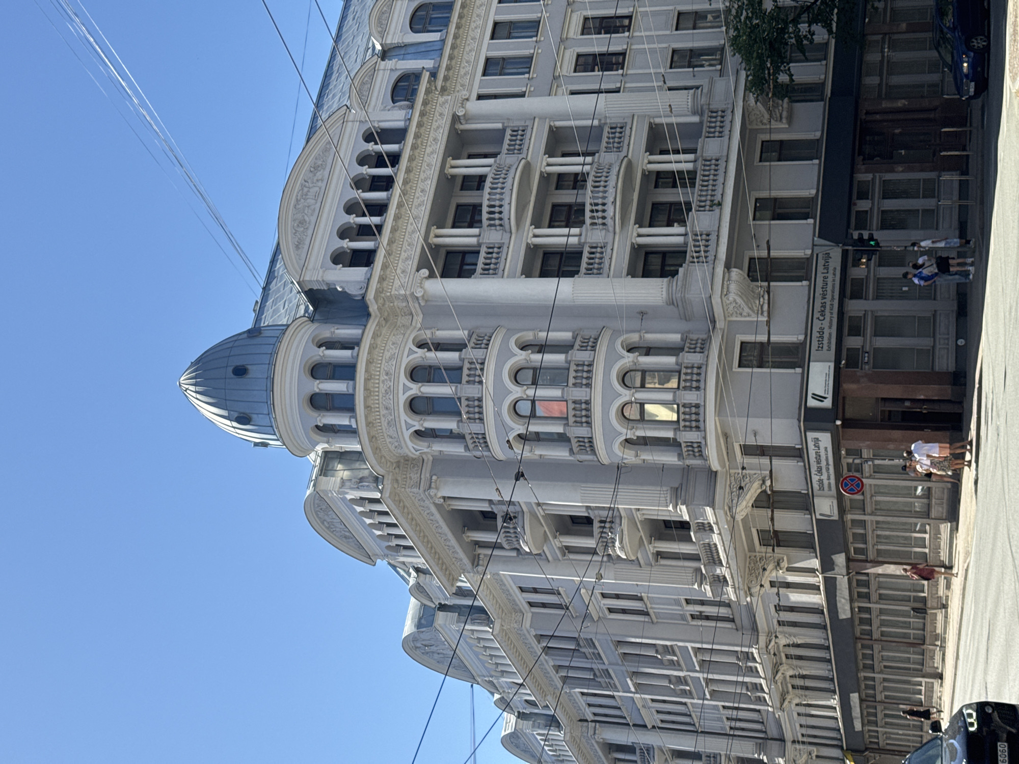This media file depicts a street view in Riga, Latvia, under a clear blue sky, suggesting a daytime scene with good weather. The primary subject is a grand, ornate building with classical architectural elements, characterized by its light-colored facade, multiple balconies with decorative railings, arched windows, and a prominent dome-like structure. The building appears to be several stories high and exhibits intricate detailing on its exterior.
In the foreground, a street is visible, with a few people standing on the sidewalk. One group of individuals is gathered near the building, appearing to be tourists or passersby. Another person is walking along the street. A dark-colored vehicle is partially visible at the bottom right of the frame, possibly a car or a bus. Overhead, several power lines or communication cables crisscross the sky, adding a sense of urban environment.
A sign on the building provides textual information. The visible text includes "Izstāde - Čekas vēsture Latvijā" and "Exhibition - History of KGB Operations in Latvia," indicating that the building houses an exhibition related to the history of KGB operations in Latvia. The presence of this sign, along with the architectural style of the building, firmly places the scene in Riga, Latvia, a city known for its historical architecture and its past under Soviet rule. The overall impression is one of a historically significant urban setting on a bright, clear day.