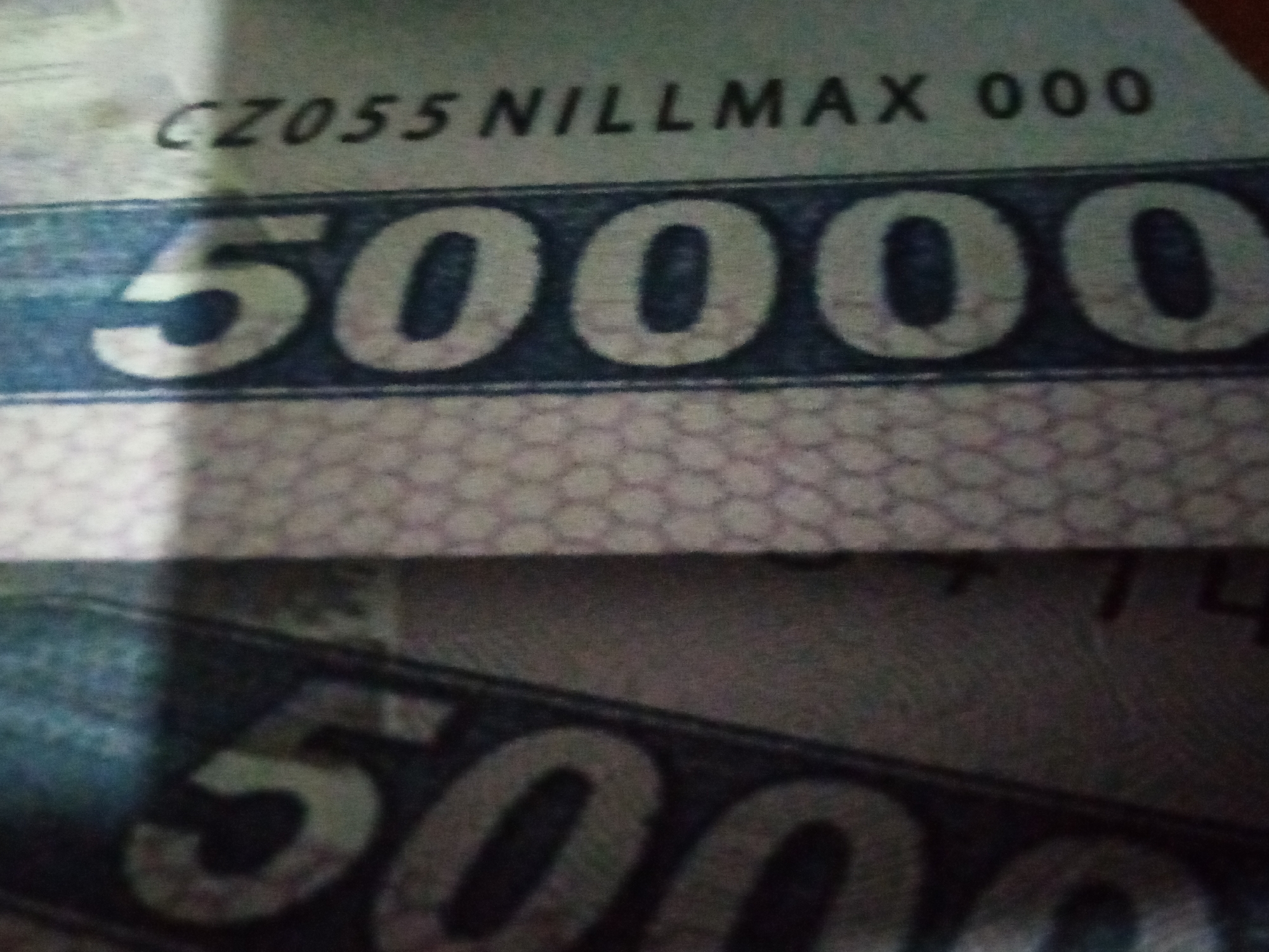 The image shows a close-up of what appears to be a banknote or a similar document. The primary visible element is the large numeral "50000" printed in a bold, white font against a dark blue background, which is itself set against a patterned beige section. Above the large numerals, there is a line of smaller text. The visible portion of this text reads "CZ055 NILLMAX 000". The image is somewhat blurry, suggesting it might have been taken with a mobile phone camera. The lighting is uneven, with a shadow obscuring the left side of the top section of the banknote. The bottom part of the image shows another, similar numeral "50000" partially visible, indicating there may be multiple notes or a larger design featuring this number. The background pattern on the beige section is a small, repeating hexagonal design. No people are visible, and the context is limited to the textual and numerical information on the document. The location context provided is Chirakchi, Uzbekistan, which might imply the banknote is of Uzbek currency, though this is not explicitly stated in the image.