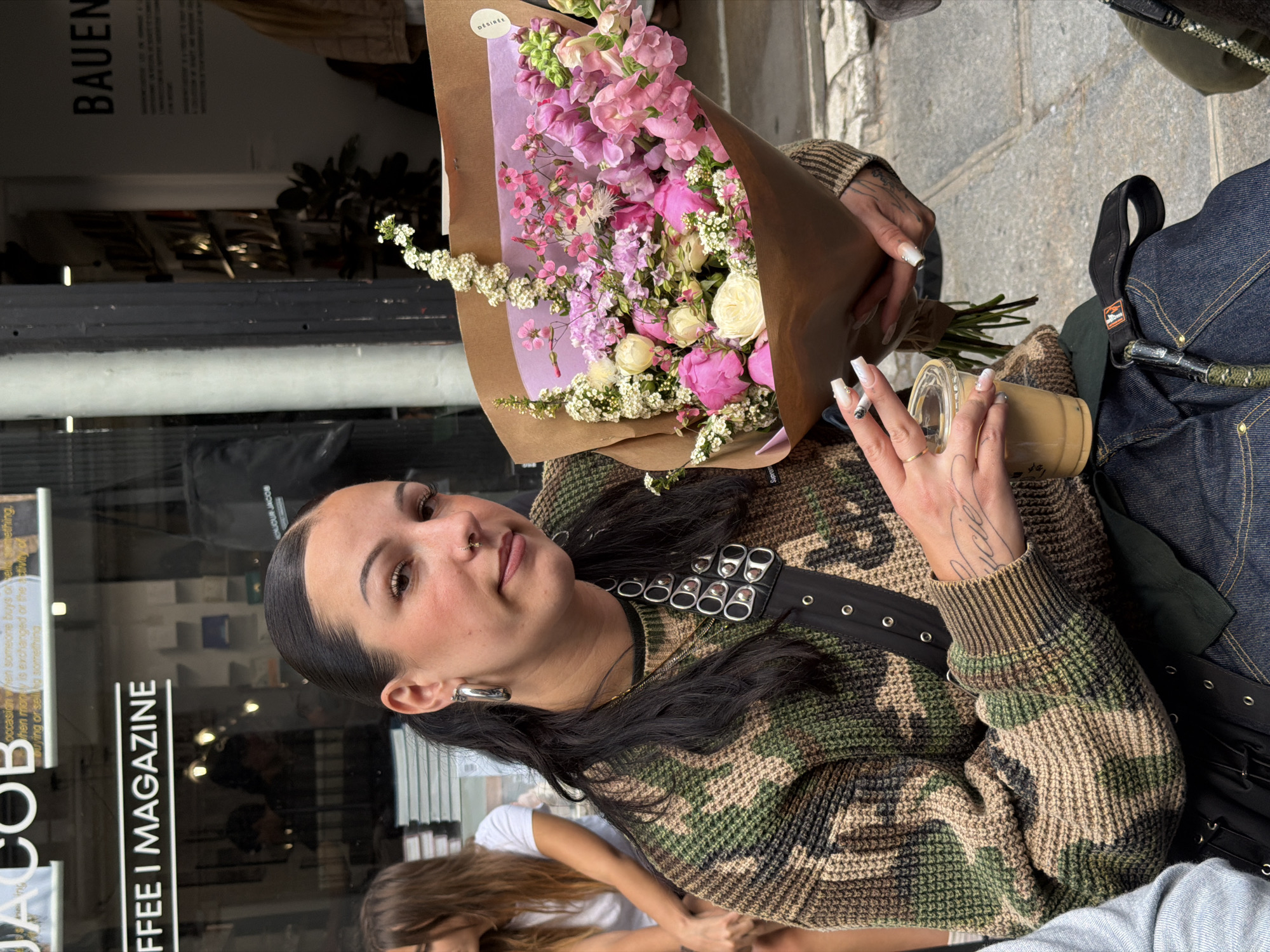 A woman with a nose ring, earrings, and tattoos is holding a bouquet of flowers in one hand and a cup of coffee in the other, smoking a cigarette. She is wearing a camouflage sweater with a studded belt and is seated outside. In the background is a storefront, with the name "JACOB COFFEE I MAGAZINE" prominently displayed. The word "BAUEN" is also visible on a sign. Another person is partially visible in the bottom left. The street looks like it could be in Paris, France.