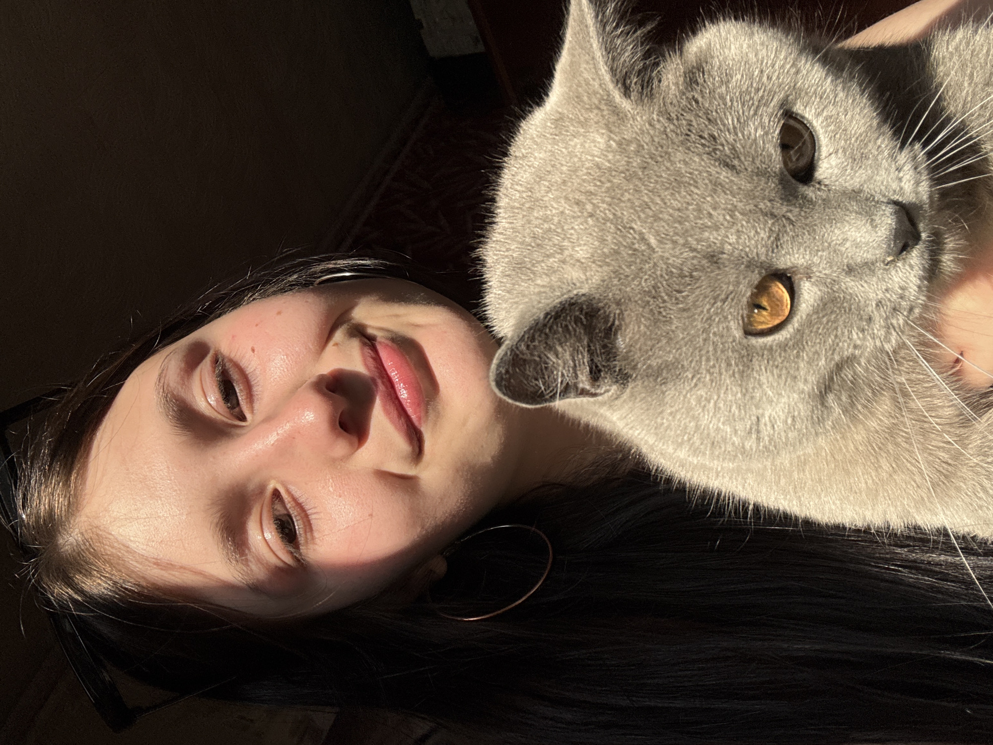 A person with fair skin, dark hair, and a hoop earring is positioned near the top-left corner of the image. The person is lying down, with their face angled towards the camera. A gray cat with golden eyes is nuzzling against the person's head. The environment is dimly lit, with the background appearing dark. The location is unknown; there is no indication the location is in Фоки, Russia.
