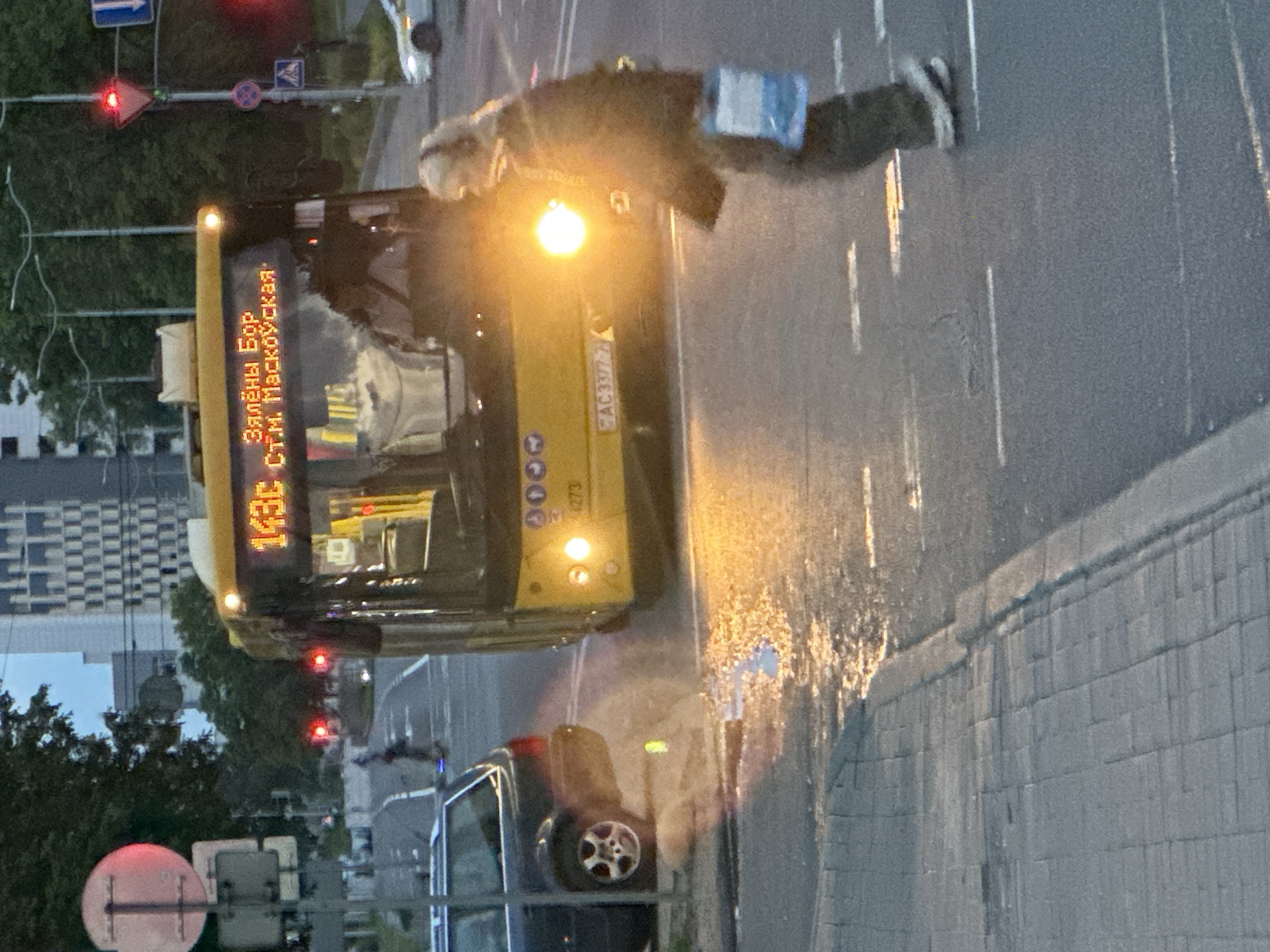 The image depicts an urban street scene in Minsk, Belarus, at what appears to be dusk or early evening, characterized by artificial illumination from streetlights, vehicle headlights, and traffic signals. In the foreground, a prominent yellow city bus occupies the center-left of the frame, facing the viewer. Its destination display shows "143c" in orange digital characters, with "Зялёны Бор" (Zialiony Bor) and "СТ.М. Маскоўская" (ST.M. Maskoŭskaja) in Cyrillic, identifying its route as connecting to the Maskoŭskaja metro station, a specific location in Minsk. The bus's headlights are brightly illuminated, casting a significant glare forward. A Belarusian license plate "АС 3377-7" is visible below the windshield, further confirming the location, with '7' indicating the Minsk region. Below the windshield and above the grille, the number "4273" is present. A pedestrian is positioned in the lower right-center, walking from right to left across the road, directly in front of the bus. The person has light-colored hair, is wearing headphones, a dark jacket or coat, dark trousers, and light-colored sneakers. They are carrying a blue and white bag in their right hand. Their facial features are somewhat obscured by the glare from the bus's headlights. The asphalt road surface shows distinct white lane markings. A significant wet patch, reflecting the bus headlights and other ambient light sources, is present on the road in the immediate foreground, suggesting recent rain or standing water. A raised curb defines the edge of a paved sidewalk on the left, constructed from rectangular pavers. In the background, multiple red traffic lights are visible, indicating that traffic may be stopped at an intersection. Various street signs are present: a large circular red and white sign (possibly prohibitory) and a smaller rectangular white sign are on a pole to the left, partially obscured by a dark-colored station wagon parked or moving slowly. This car's rear left wheel and a portion of its rear bumper are visible. To the right, further back, a red triangular "yield" sign, a circular red and white sign (possibly prohibitory), and a blue square sign with a white pedestrian icon (indicating a pedestrian crossing) are visible. A white car, possibly a taxi, is in the far right background. Trees line both sides of the road, and in the distant background, multi-story buildings with a modern architectural style and grid-patterned windows are visible, characteristic of an urban environment. The sky appears overcast or dark, consistent with dusk