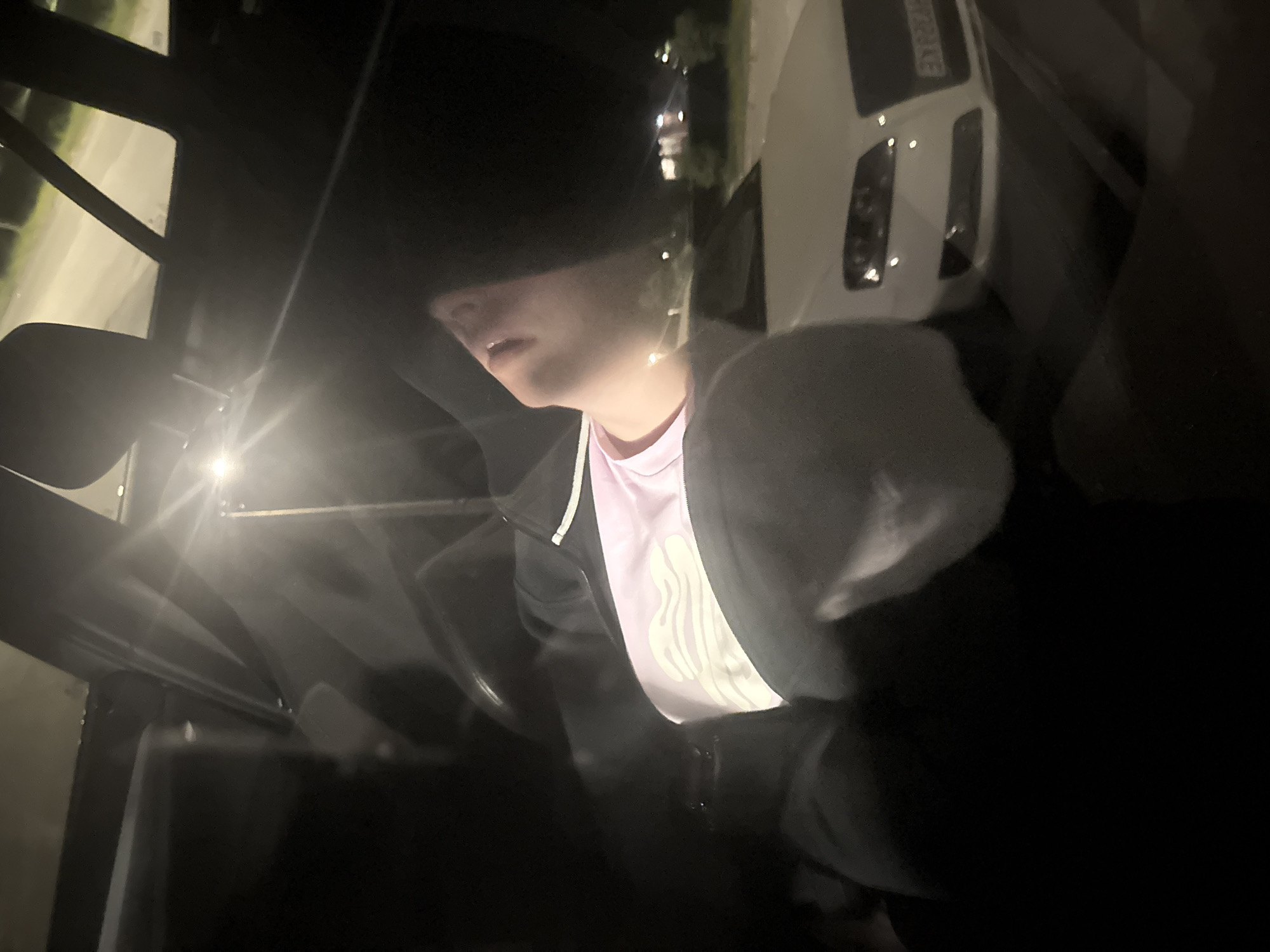 This nighttime photograph, taken in Kyustendil, Bulgaria, depicts the interior of a vehicle and an exterior street scene. Inside the vehicle, a person, oriented in a reclining or supine position, is visible. They are wearing a black hooded jacket over a light pink garment with a white, wavy pattern across the chest. Their face, partially obscured by shadow, is angled upwards. A strong external light source, likely a streetlamp, creates a significant lens flare originating from the upper right portion of the image. Through the car's window, a white Audi sedan is parked, with its front end, headlights, and a partially visible license plate ("EX 0284" or similar) discernible. The "EX" prefix on the license plate corresponds to the Kyustendil registration region. The background features a street, distant streetlights, and dimly lit structures, confirming an outdoor urban or suburban environment.