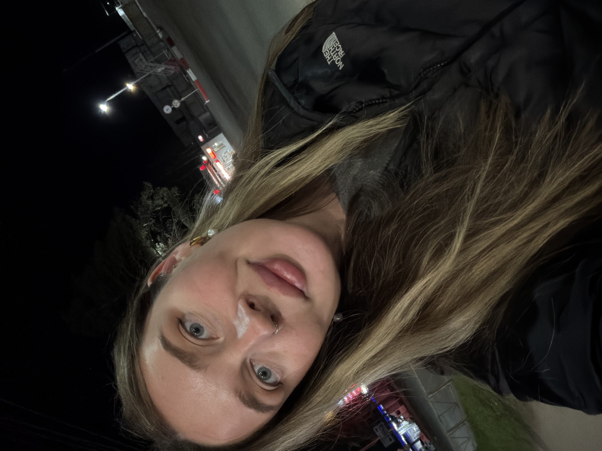 A woman with light-colored hair is taking a selfie at night. She has blue eyes, fair skin, and a nose ring. She is wearing a black North Face jacket. There is a patch of white cream on her face, near her nose. In the background, there are street lights and a building with illuminated signs. The photo appears to be taken in an urban setting in the city of Chaikovsky, Russia.