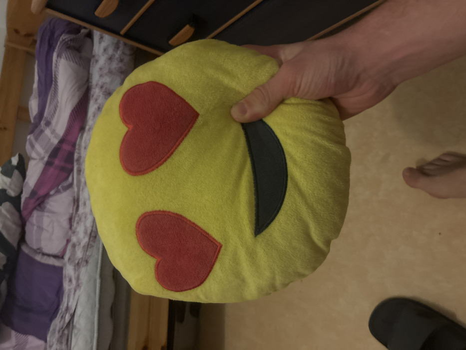 A hand holds a yellow, round, plush pillow resembling a love-struck emoji. The pillow features two red heart shapes for eyes and a curved, black smile. A partially visible foot and a black slipper sit on a tan-colored surface. In the background are a wooden dresser and patterned bedding. The scene is well-lit, highlighting the textures of the pillow and the surrounding elements.