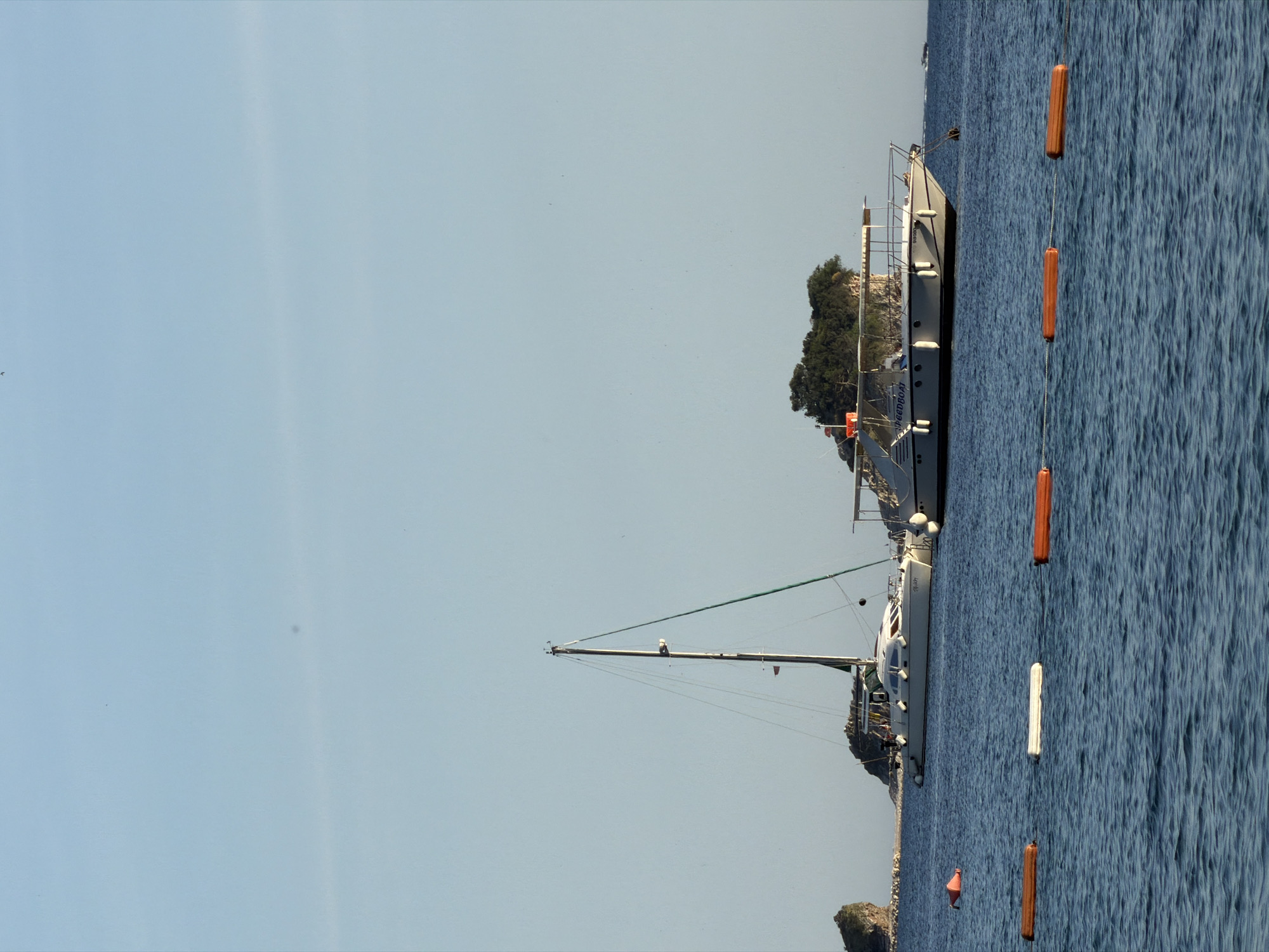 The image depicts a coastal scene in Budva, Montenegro, featuring a large yacht anchored in the clear blue water. The yacht is positioned near a small, tree-covered island or peninsula. A line of buoys is visible in the water, likely marking a swimming area or channel. The sky is clear and blue, suggesting a sunny day. The orientation of the image is vertical, presenting the scene from a slightly off-kilter perspective.