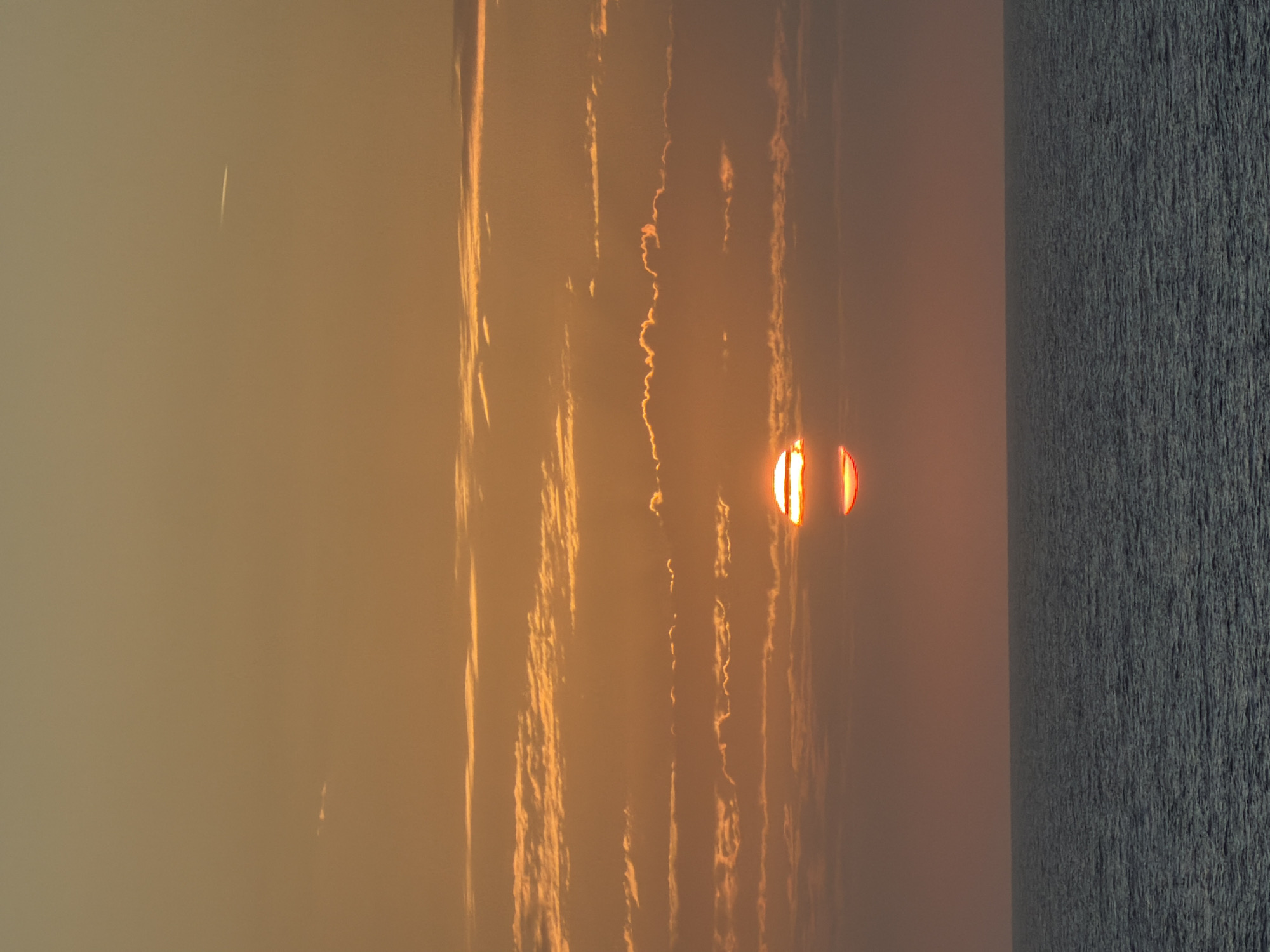 The image depicts a sunrise or sunset over a body of water, captured with a warm, golden-orange color palette. The scene is dominated by a hazy sky and textured water, creating a tranquil atmosphere.
The sun, positioned low on the horizon towards the right, appears as two distinct, intensely bright orange-red spheres, suggesting atmospheric distortion, a strong lens flare, or a reflection. Its light illuminates long, vertical (when correctly oriented) cloud formations stretching across the sky, painting them in hues of bright orange, yellow, and deep gold. Some clouds are thin and wispy, while others form more substantial, almost painterly streaks of light against the muted background sky. A faint, thin white streak, possibly a distant airplane contrail, is visible in the upper-left portion of the sky.
The lower part of the image is occupied by a dark, textured body of water, likely an ocean or a large lake, which reflects very little of the vibrant sky, appearing somewhat dark and choppy. The overall impression is one of natural grandeur and a serene, early morning or late evening moment. There is no visible text in the image.