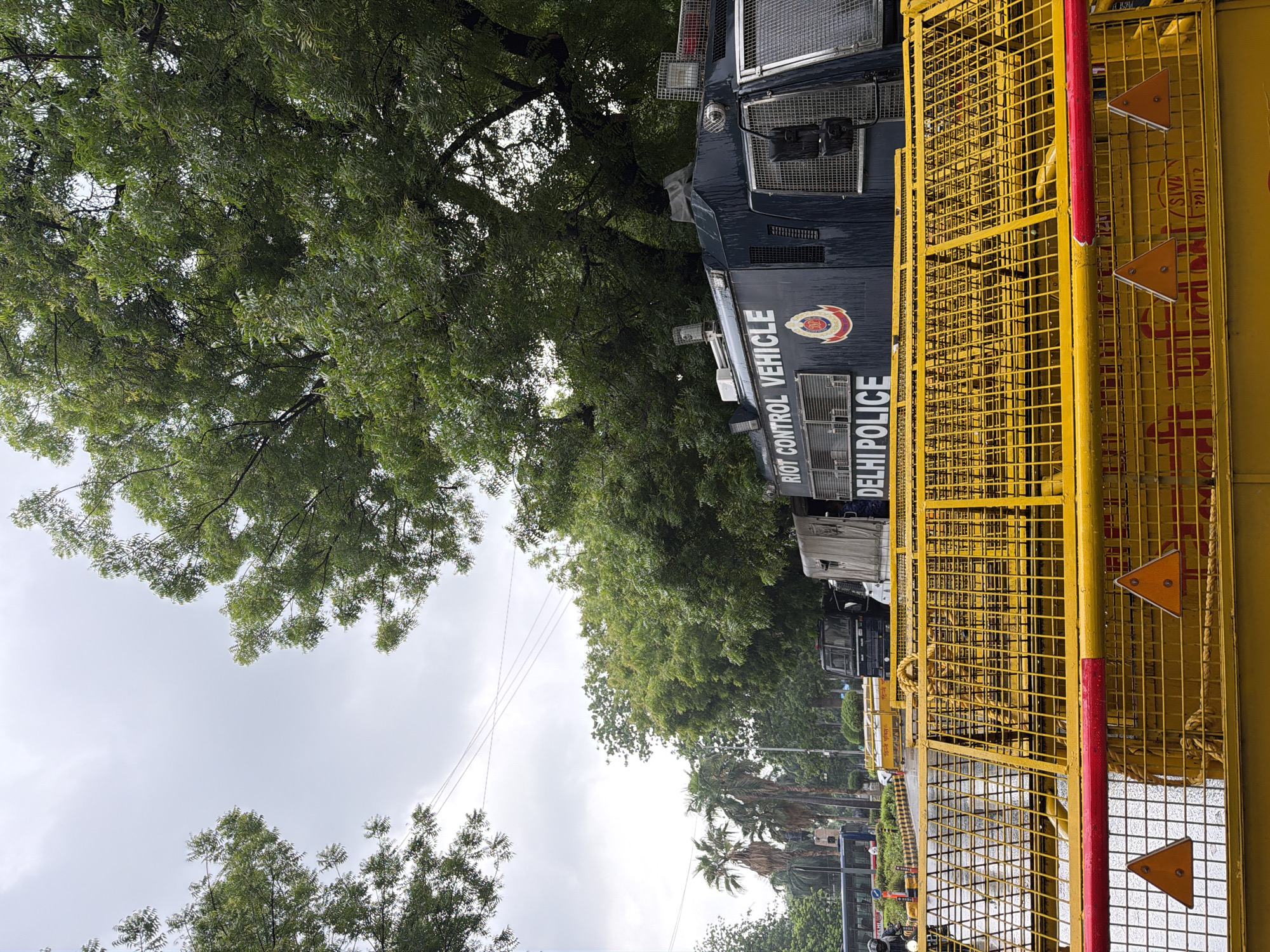 The image depicts a street scene in New Delhi, India, with a riot control vehicle and barricades prominently featured. A dark blue riot control vehicle, identified by the text "RIOT CONTROL VEHICLE" and "DELHI POLICE" on its side, is parked behind a bright yellow metal barricade. The barricade is composed of multiple sections and includes red markings and orange triangular reflectors. In the background, trees with lush green foliage line the street, and a cloudy sky suggests overcast weather. Other vehicles, including a bus and what appear to be police trucks, are visible further down the road. The presence of the riot control vehicle and barricades indicates a security presence, possibly in response to a planned event or ongoing situation. The overall environment appears to be an urban street within New Delhi.