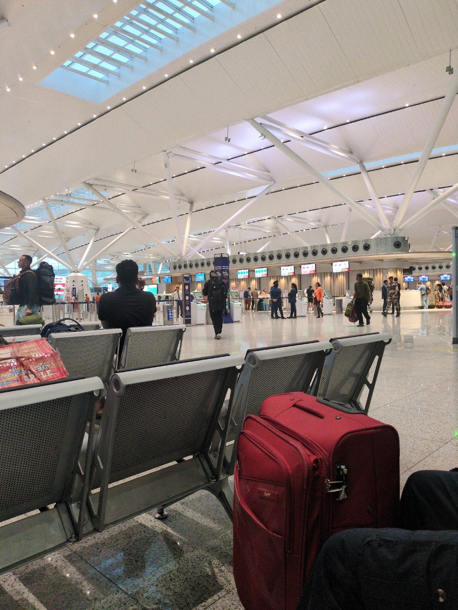 The image shows the interior of a modern airport terminal with a high, vaulted ceiling and large windows. In the foreground, there are rows of empty metal airport seats, with a red suitcase and a person's dark clothing on the seats to the right. In the mid-ground, people are walking and standing, some with luggage. There are check-in counters visible in the background, with screens displaying flight information. The environment is clean and well-lit, with a polished floor reflecting the overhead lights. The scene suggests a busy travel hub.