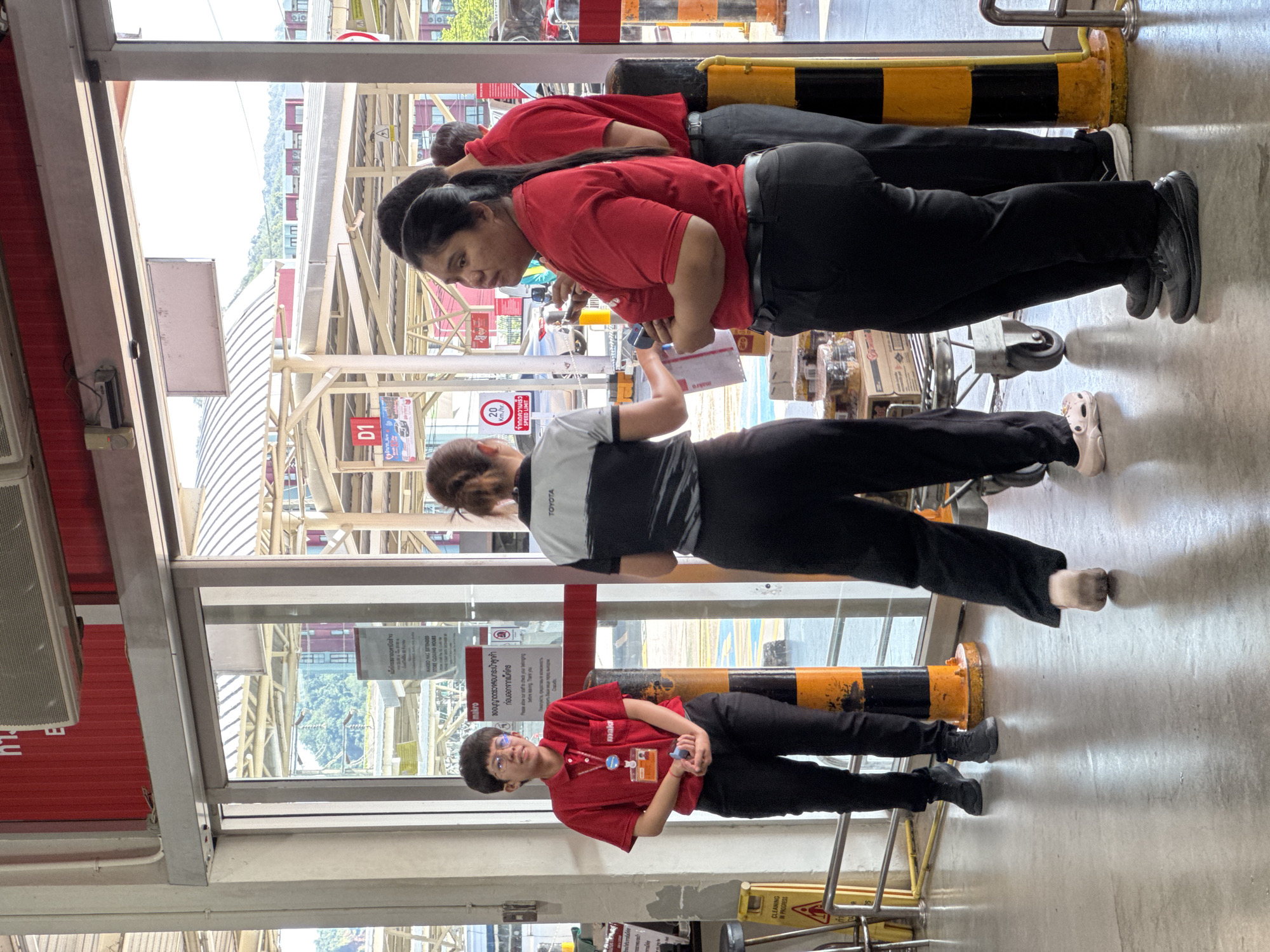 In Wichit, Thailand, near the entrance of a Makro store, four individuals are standing on a concrete floor. A woman wearing a red polo shirt and black pants, with glasses and short hair, stands near a glass door holding a blue item. To her right, another woman in a black and gray collared shirt with "TOYOTA" on the sleeve and black pants faces away from the camera. To the right of her, a woman in a red polo shirt and black pants looks towards the glass door. Next to her, a man in a red polo shirt and black pants stands facing the door. In the background, a shopping cart filled with merchandise is visible, along with signage and the exterior of the building.