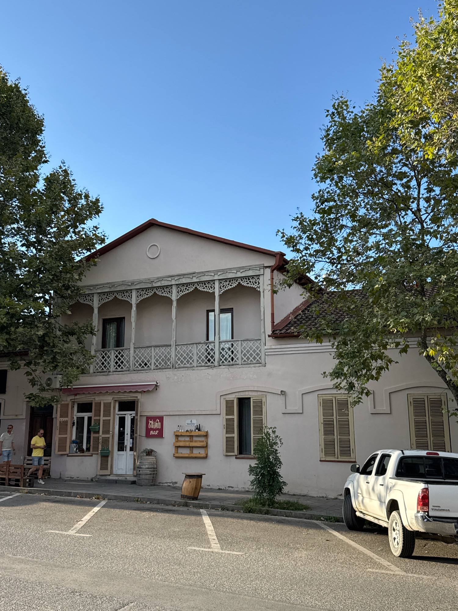 The image shows a street scene in Telavi, Georgia. A light-colored, two-story building with a decorative wooden balcony on the upper floor is the main subject. The building has several windows with wooden shutters. A sign above a ground-floor entrance reads "bar". A wooden wine rack with bottles is mounted on the exterior wall, and two wooden barrels are placed near the entrance. In front of the building, there is a sidewalk and a paved street with white parking lines. A white pickup truck is parked on the right side of the street. Two men are standing on the sidewalk to the left of the building. Trees with green foliage are visible on the left and right sides of the image. The sky is clear and blue.