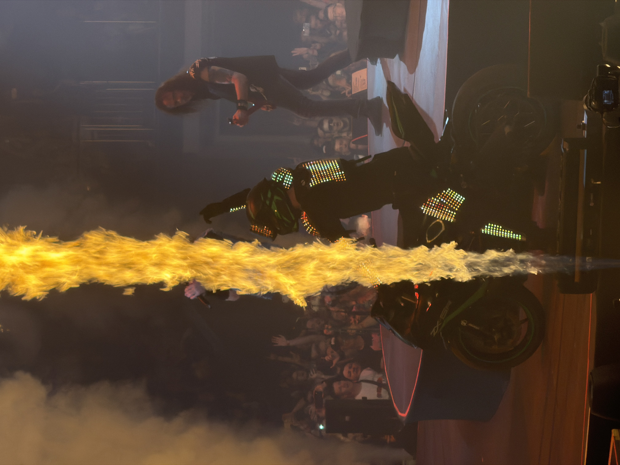 A concert performance is captured, featuring a person in a black costume adorned with colorful LED lights, riding a black and green motorcycle. The motorcycle is positioned on a stage with a red-lit circular platform. A column of fire erupts vertically from behind the motorcycle, obscuring a portion of the performer. In the background, a guitarist with long dark hair, wearing a black sleeveless shirt, plays a guitar. A crowd of concertgoers is visible behind the performers. The environment appears to be an indoor venue, likely a concert hall, with stage lighting and smoke effects. The date and specific location of the concert in Saint Petersburg, Russia are not discernible from the image alone.
