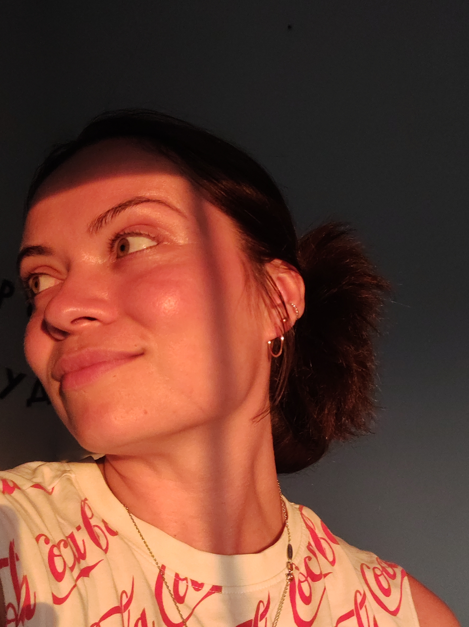 The image shows a close-up portrait of a young woman with dark hair pulled back in a bun. She is wearing a light-colored t-shirt with a repeating red Coca-Cola logo pattern and a delicate gold chain necklace. The woman is looking to her right, with a slight smile. A strong shaft of light casts a shadow across her face, highlighting her features. The background is a plain, dark gray wall. The image does not contain any information that could be used to determine the specific location in the city of Foky, Russia.