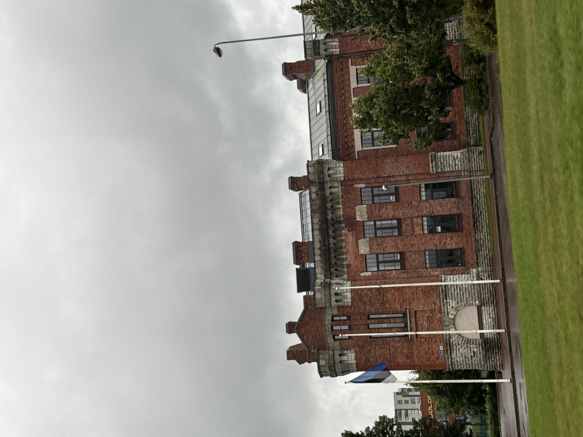The image shows the exterior of the Kalaranna Fortress in Tallinn, Estonia, under a cloudy sky. The fortress is a red brick building with grey stone accents and several windows. Two flagpoles with the Estonian flag are visible in front of the building. There is a street and a grassy area in the foreground. Trees are present on the right side of the image. In the background, another building with the word "GOLDE" is visible. A street lamp is also present on the right side of the frame. The scene appears to be a public outdoor space.